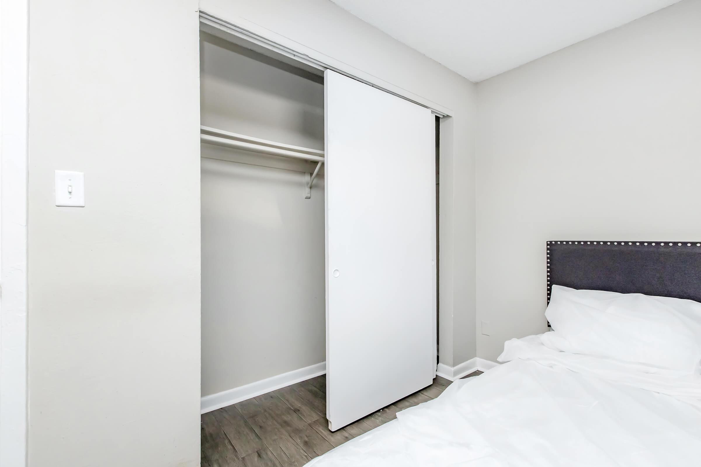 A well-lit bedroom featuring a neatly made bed with white bedding. To the side, there is an open closet with sliding white doors, revealing a simple shelving area. The walls are painted in a light color, and the flooring appears to be wood laminate.