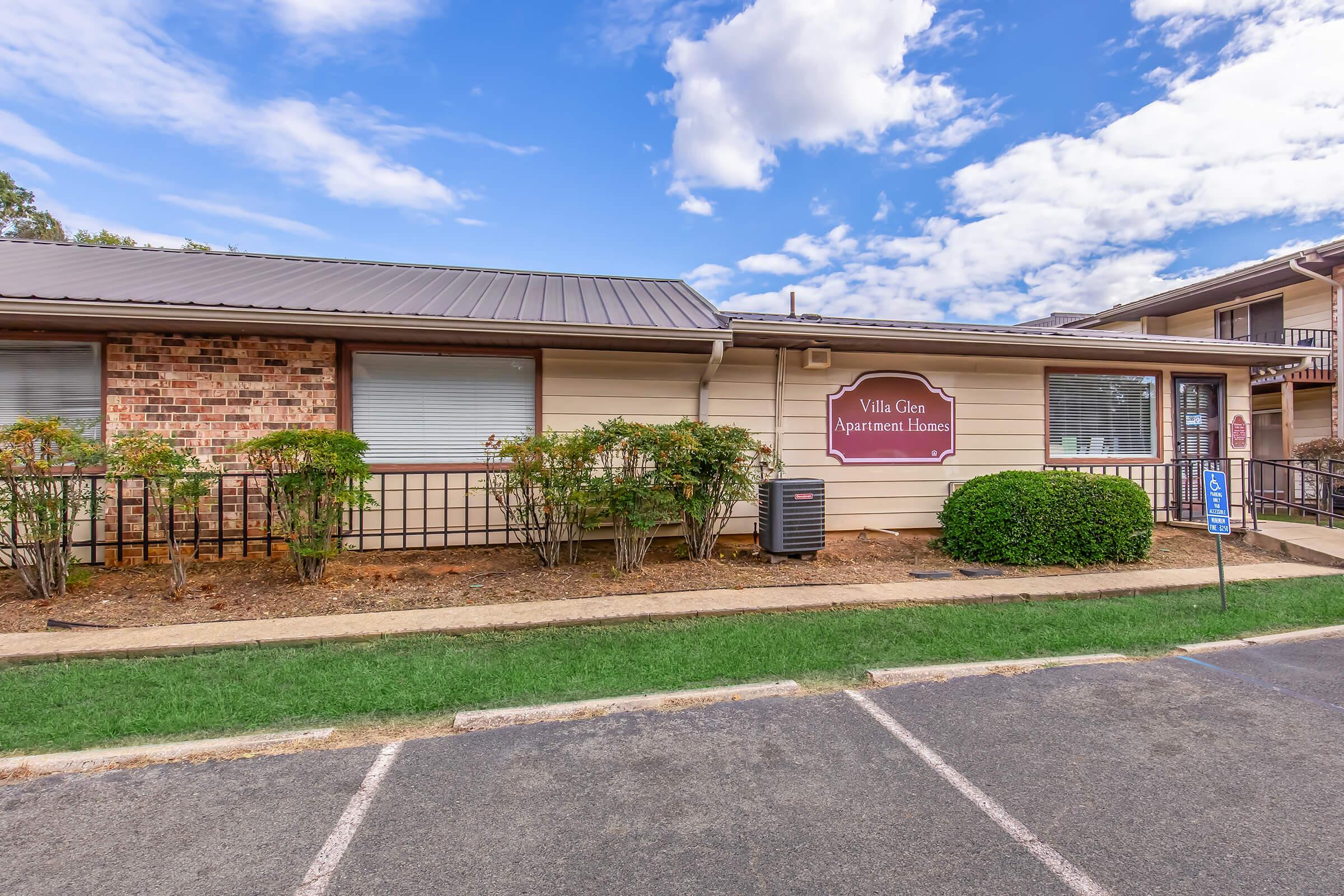 Exterior view of Villa Glen Apartment Homes, featuring a brick and siding facade under a metal roof. The building is surrounded by well-maintained landscaping, including shrubs and grass. The sky is partly cloudy, and the image includes a designated parking space nearby.