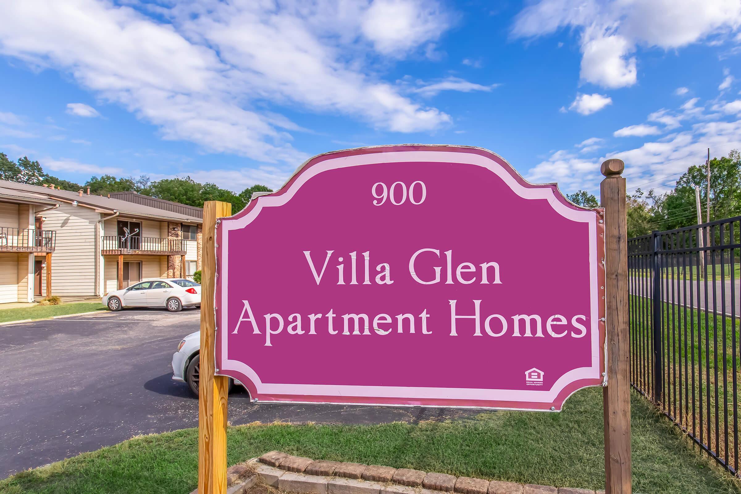 Sign displaying "Villa Glen Apartment Homes" with the address "900". The sign is pink and wooden, located near an apartment complex. In the background, there are trees and a clear blue sky, and a parking area with cars can be seen.