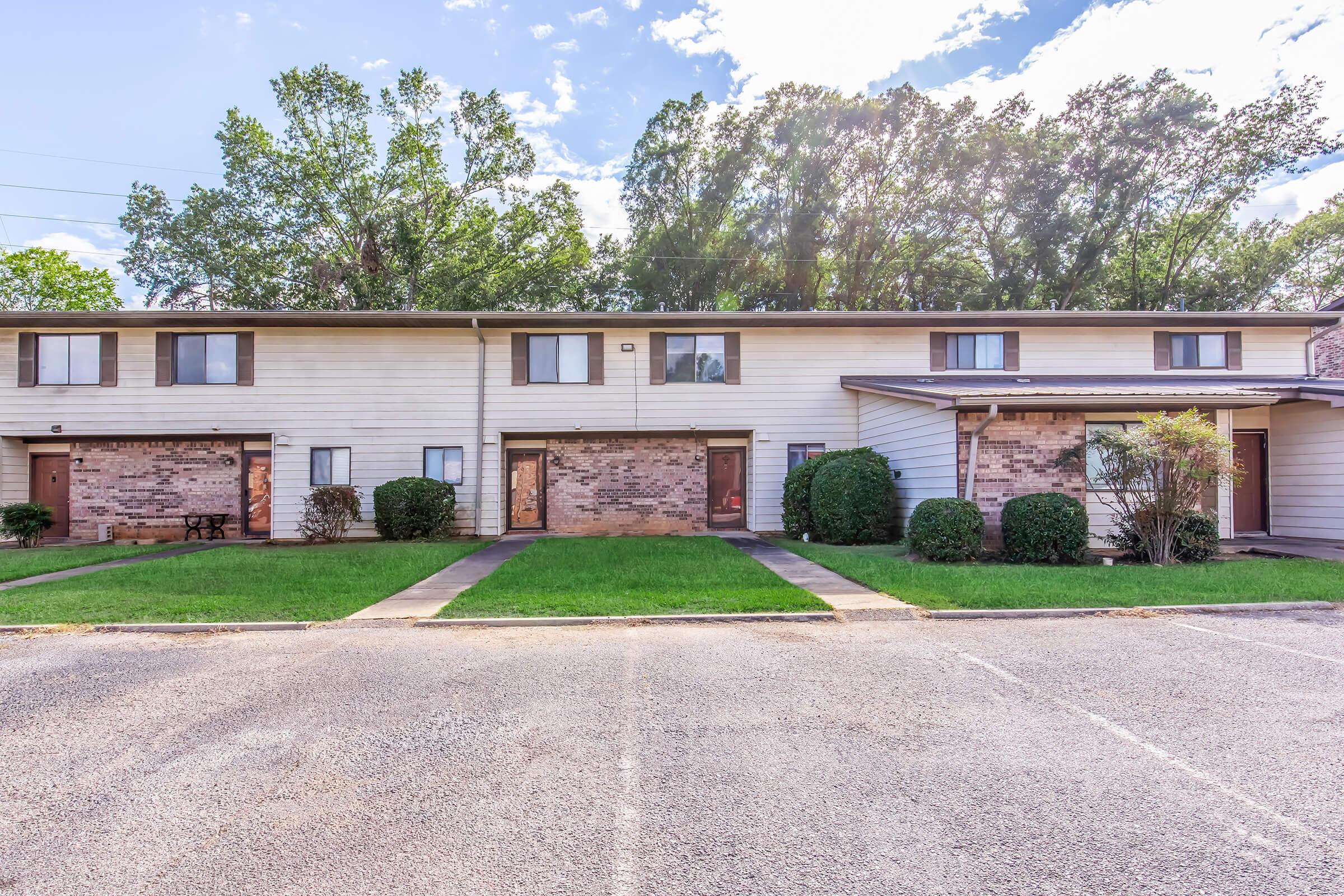 A row of two-story townhouses with brick and siding exteriors, featuring green lawns and small shrubs in front. The pathway leads to individual entrances, and the sky is partially cloudy with sunlight illuminating the scene. Parking spaces are visible in the foreground.