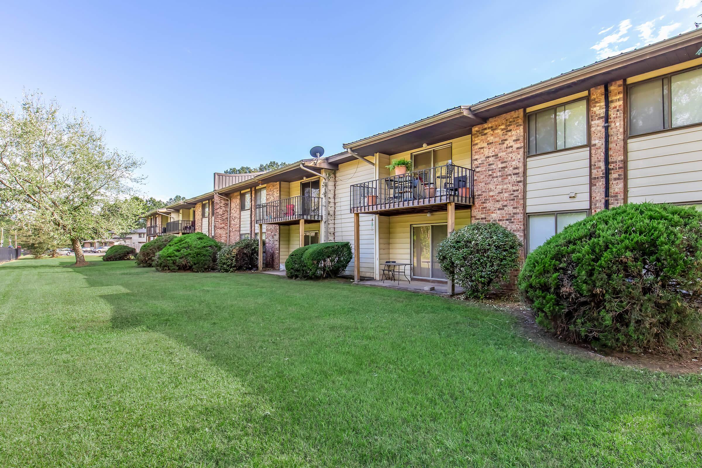 Row of apartment buildings with brick facades and balconies, surrounded by well-maintained green lawns and shrubs. Bright blue sky in the background.