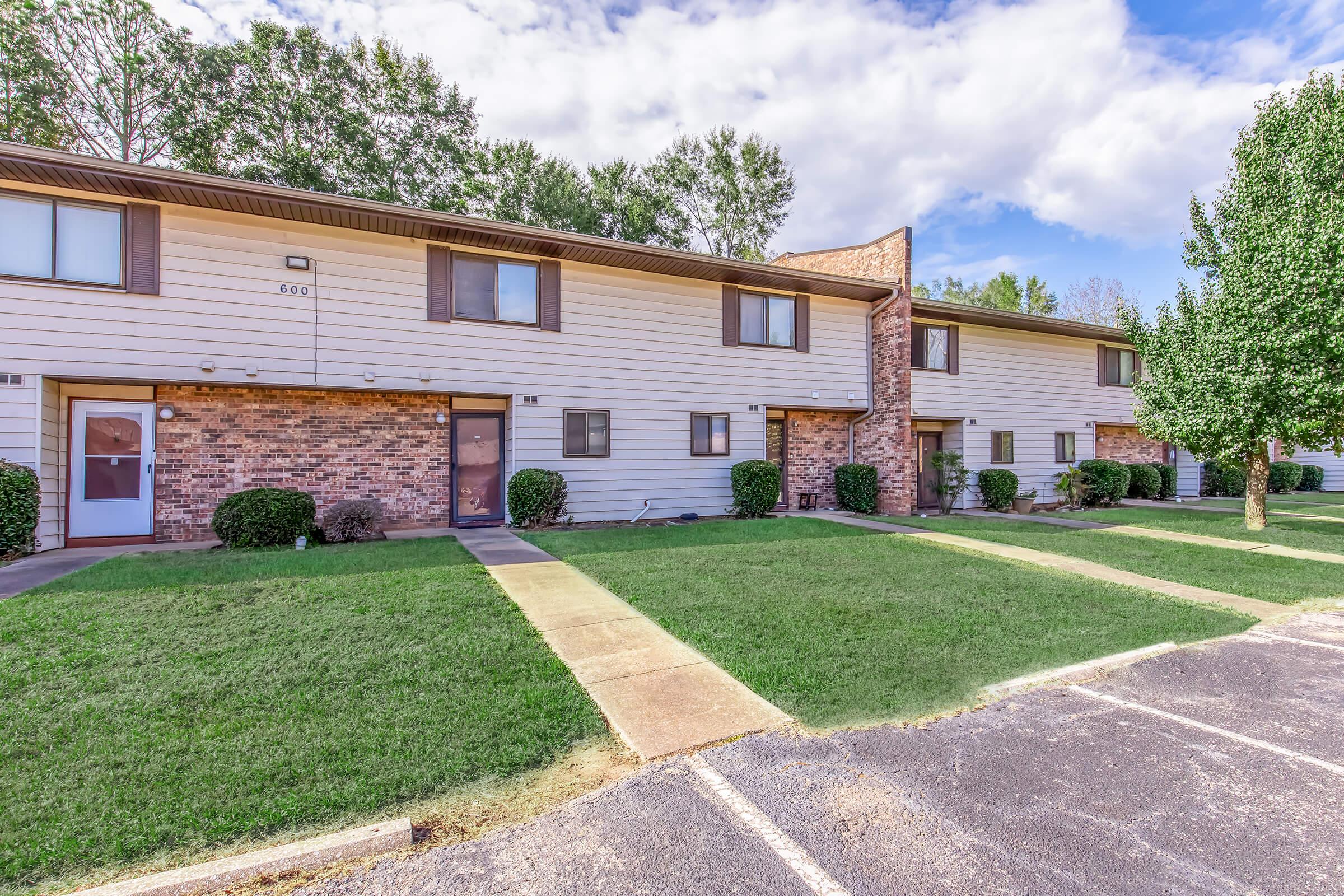 Two-story apartment complex with multiple units featuring brick and siding exteriors. Neatly trimmed lawns and small shrubs surround the buildings. A paved parking area is visible in the foreground, and trees line the side of the property under a partly cloudy sky.