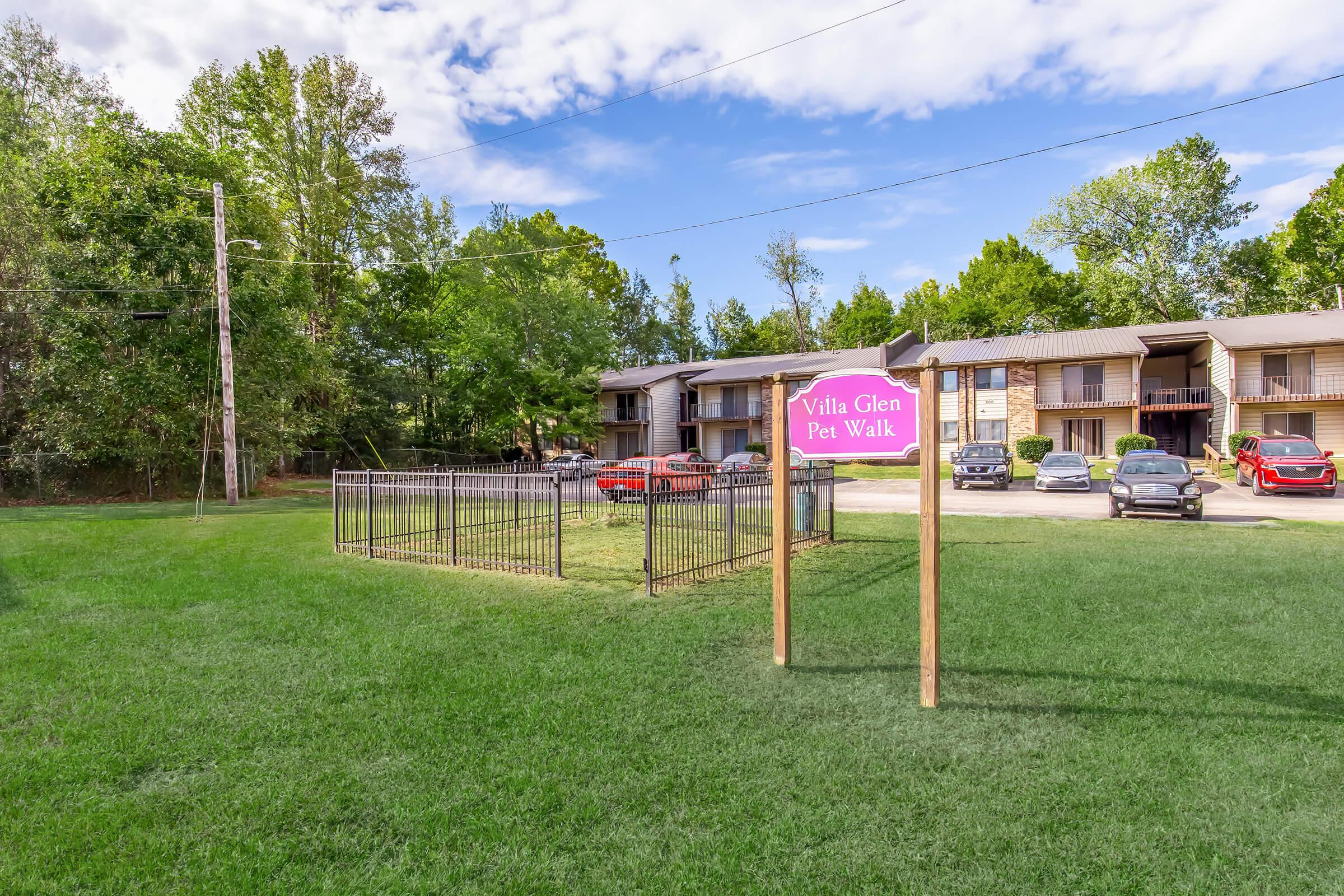 A grassy area featuring a sign that reads "Villa Glen Pet Walk." There is a fenced space for pets, with several parked cars and apartment buildings visible in the background. The scene is set under a partly cloudy blue sky with trees surrounding the area.