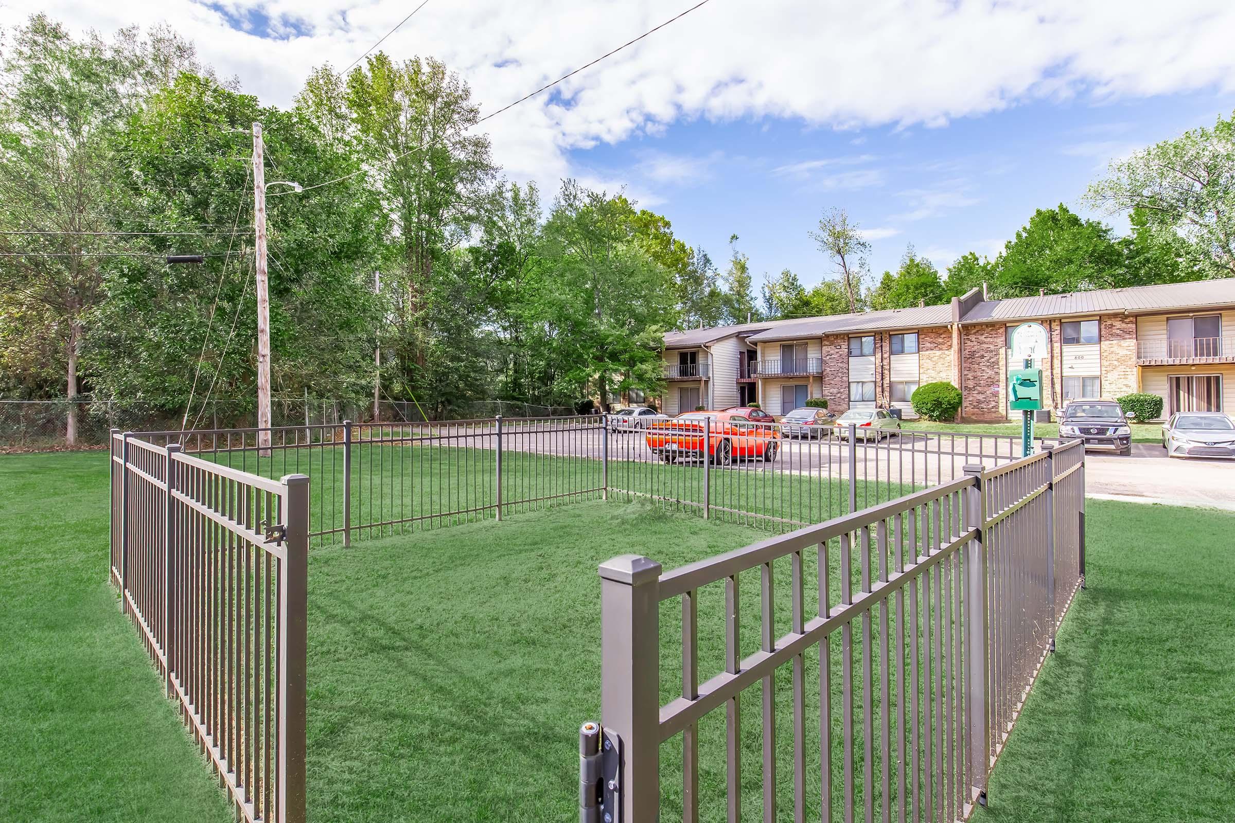 A fenced area with green grass is seen in the foreground. In the background, there are two-story apartment buildings surrounded by trees. A parking area with several cars is visible. The sky is partly cloudy, creating a bright atmosphere.
