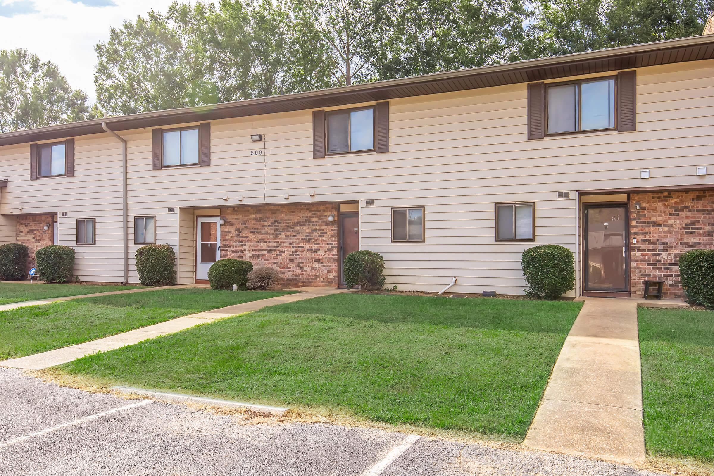 A row of beige and brown apartment units with two levels, featuring front doors with small windows. Neatly trimmed grass and shrubs line the walkways leading to each entrance. The scene is set in a suburban area with trees in the background under a clear sky.