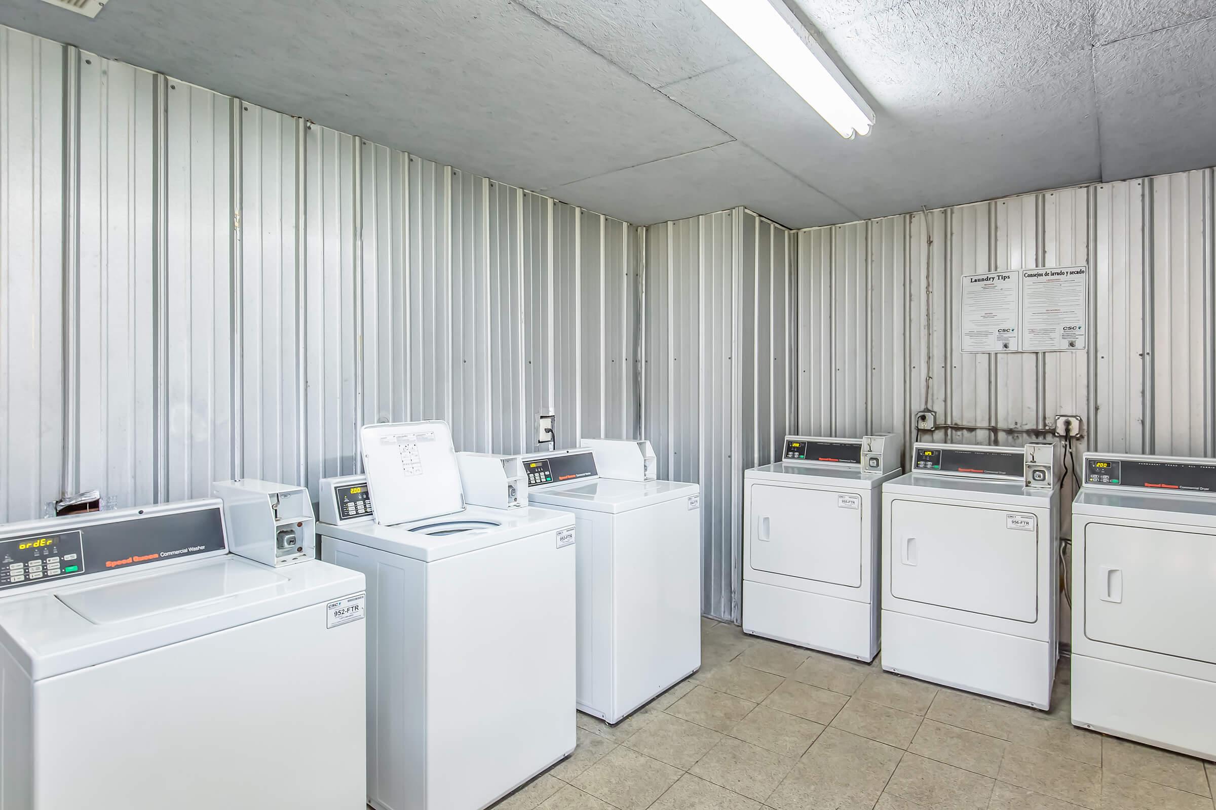 A clean and well-lit laundry room featuring multiple white washers and dryers arranged against a metallic wall. The floor is tiled, and there are overhead lights illuminating the space. A notice board is mounted on the wall, providing information about laundry usage.