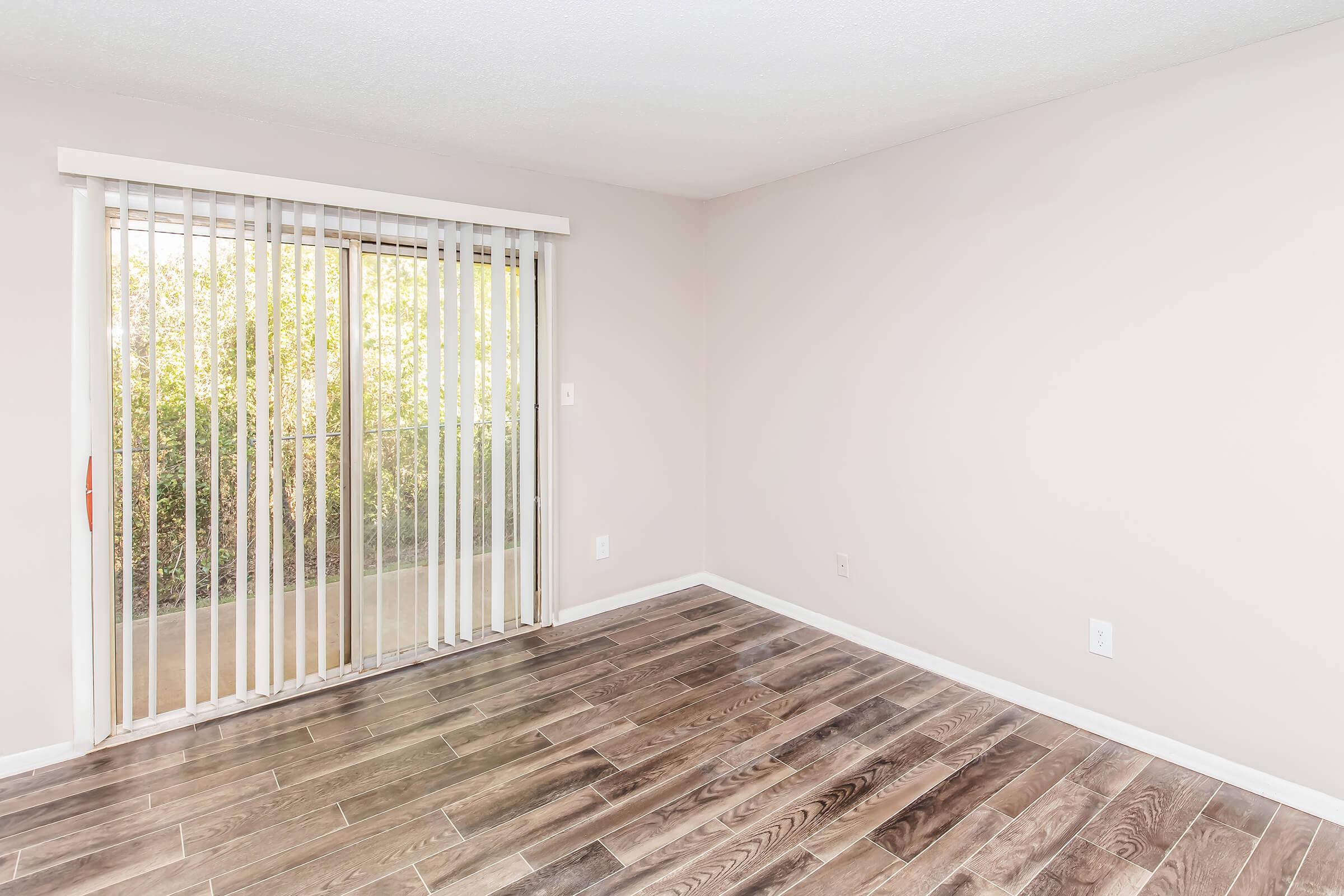 Empty interior of a room featuring light-colored walls, a sliding glass door with vertical blinds, and wooden flooring. The space appears bright and uncluttered, with natural light coming through the door, and a small view of greenery visible outside.