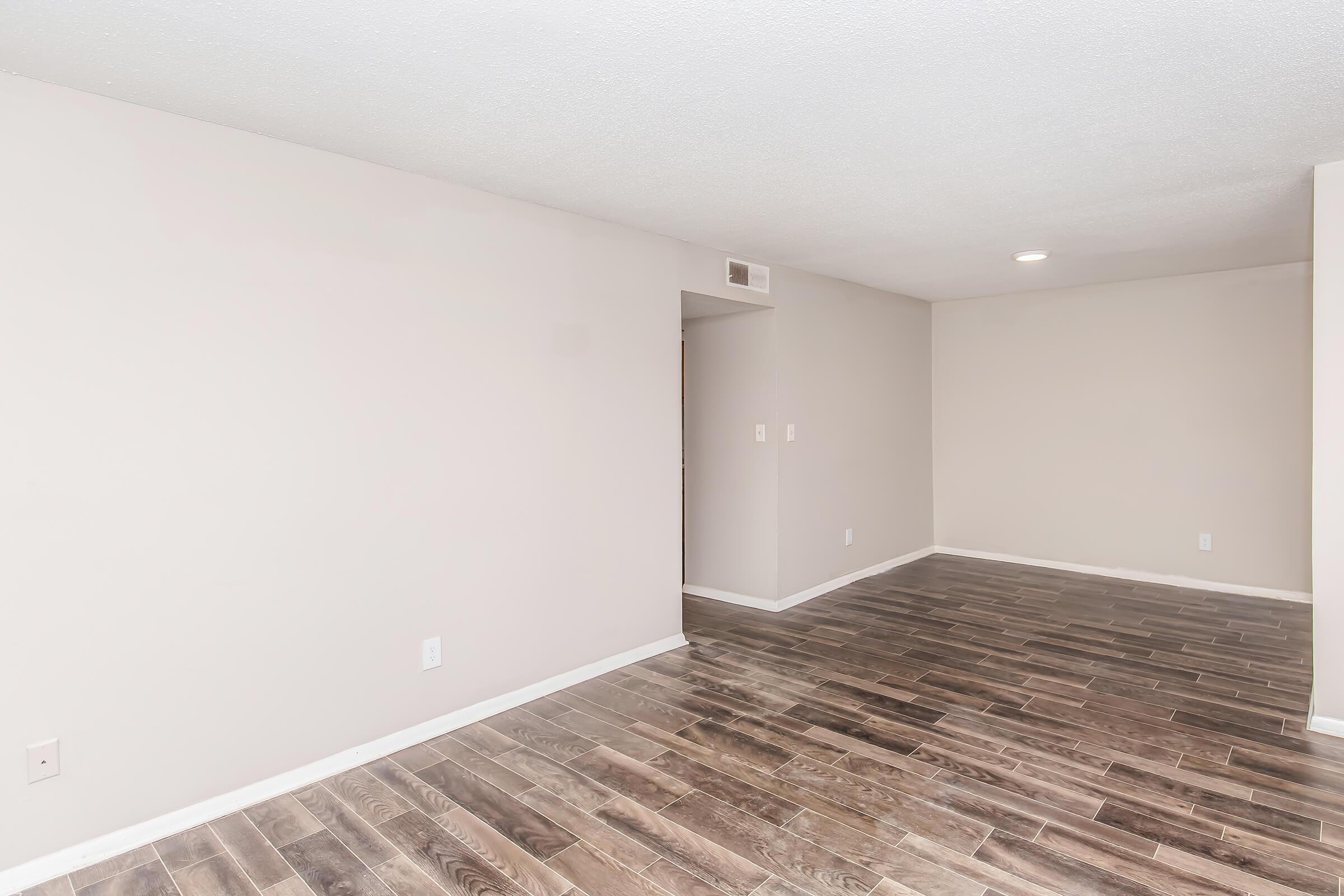 A spacious, empty room with light gray walls and dark wood-like flooring. There are no furnishings or decorations, and the lighting is soft and even. A doorway is visible on the left, leading to another area, and a ceiling light fixture is present. The overall atmosphere is bright and clean, suitable for various uses.