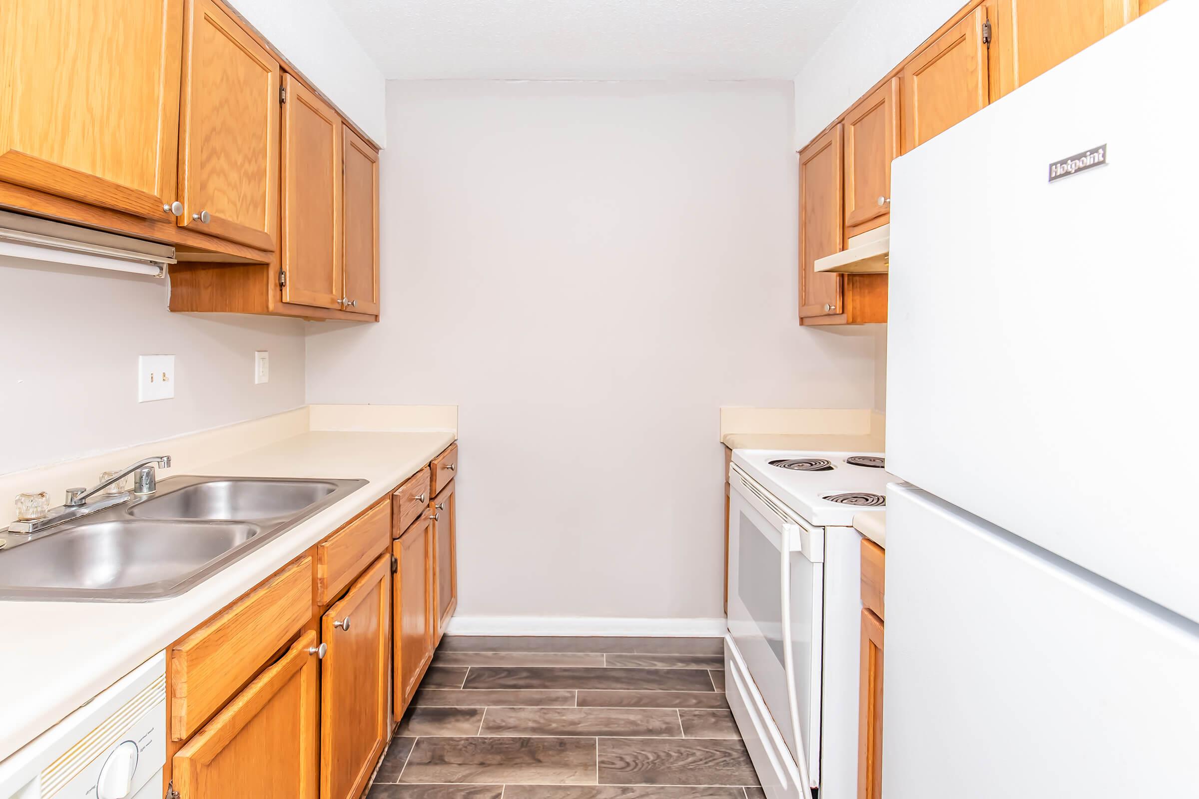 A modern kitchen featuring wooden cabinets, a double sink, a white refrigerator, and an electric stove. The walls are painted light grey, and the floor is covered with dark tiles, creating a clean and inviting space. Countertops are beige, complementing the cabinetry.