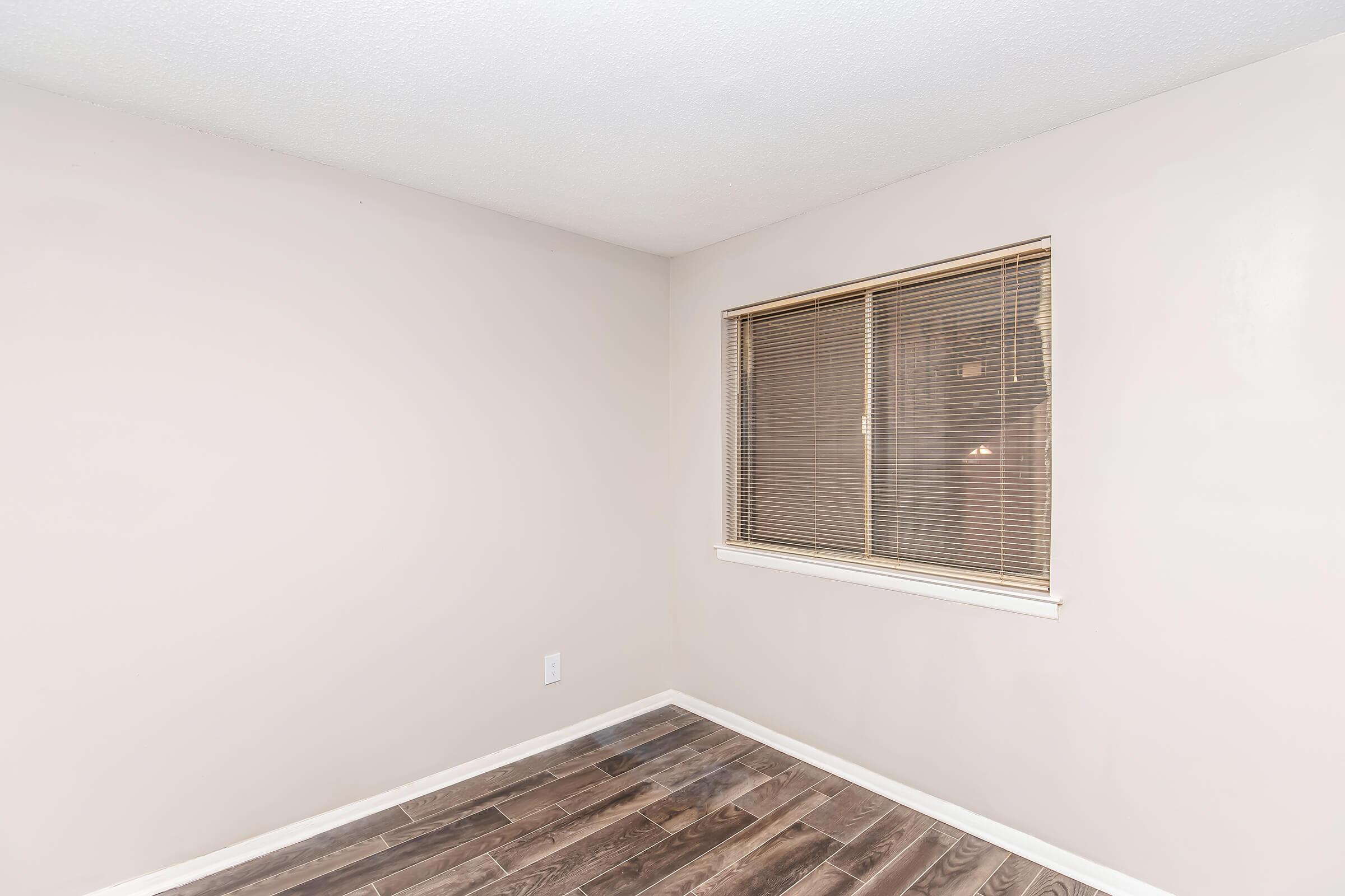 Empty room with light beige walls and a wooden floor. A window with horizontal blinds is located on one side, allowing natural light to enter the space. The room appears clean and uncluttered, creating a minimalist atmosphere.