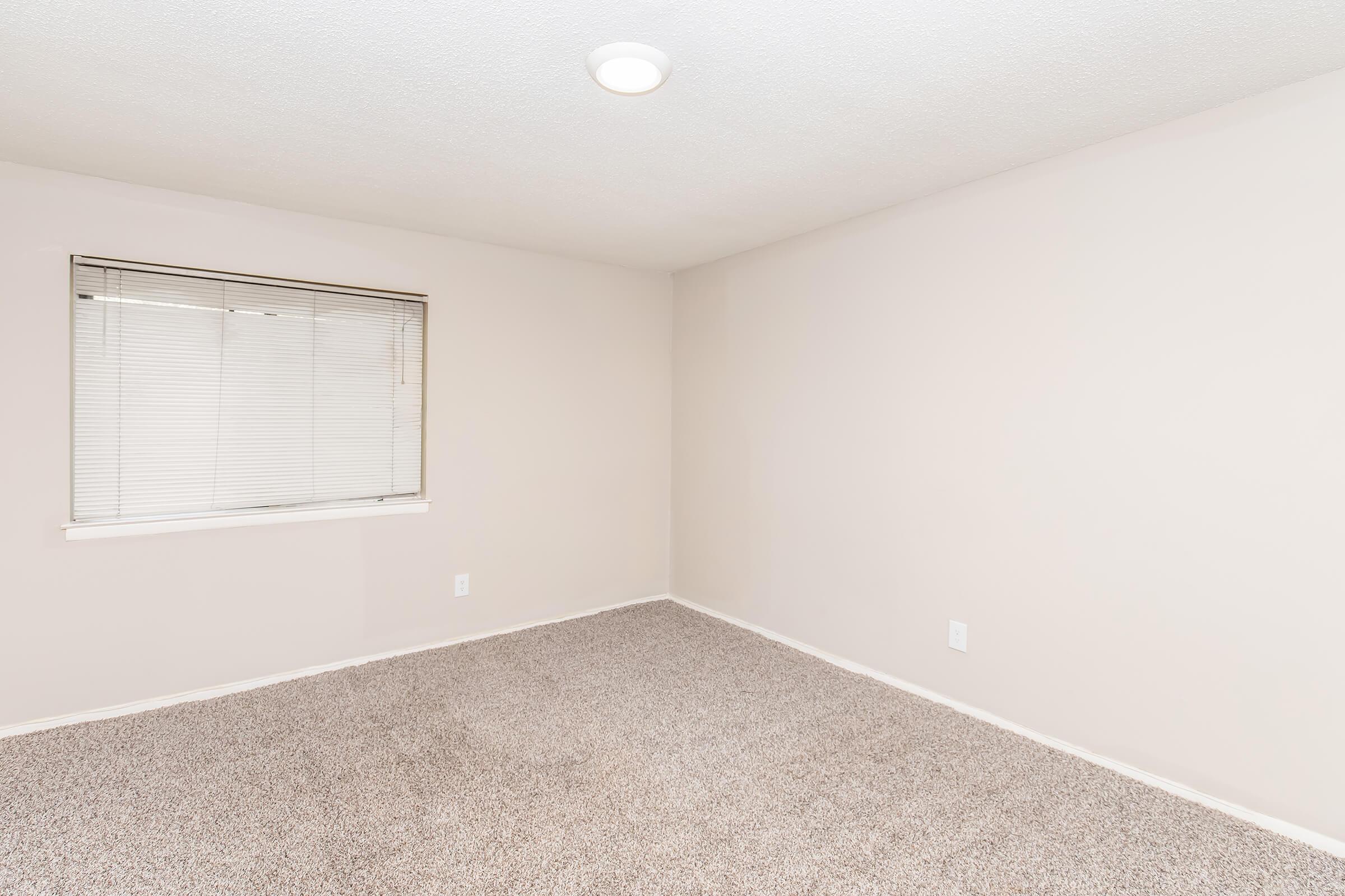 Empty room with beige walls and carpet, featuring a window covered with vertical blinds. Soft overhead lighting illuminates the space, creating a neutral and inviting atmosphere.