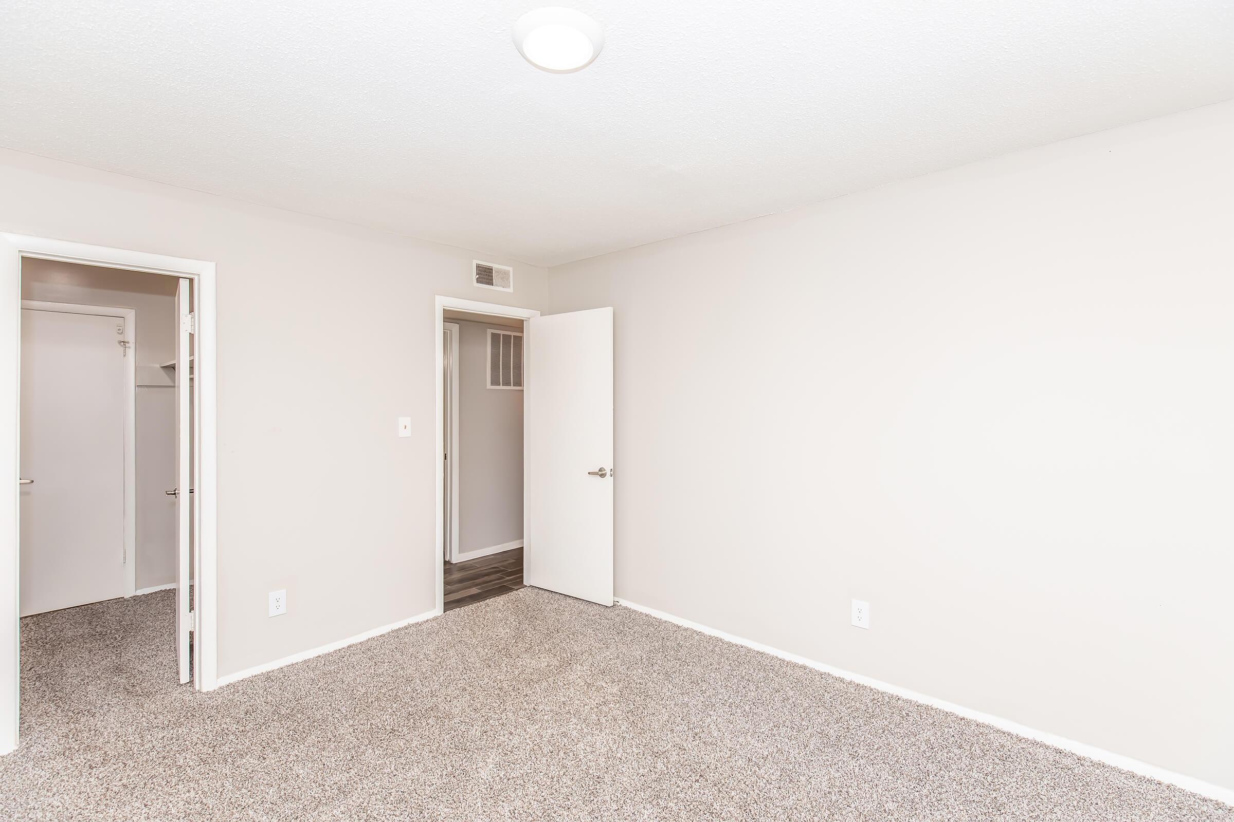 A spacious, empty room with light-colored walls and plush carpet. There are two open doorways leading to adjacent rooms, and soft overhead lighting enhances the bright atmosphere. The room is clean and ready for furniture or decoration.