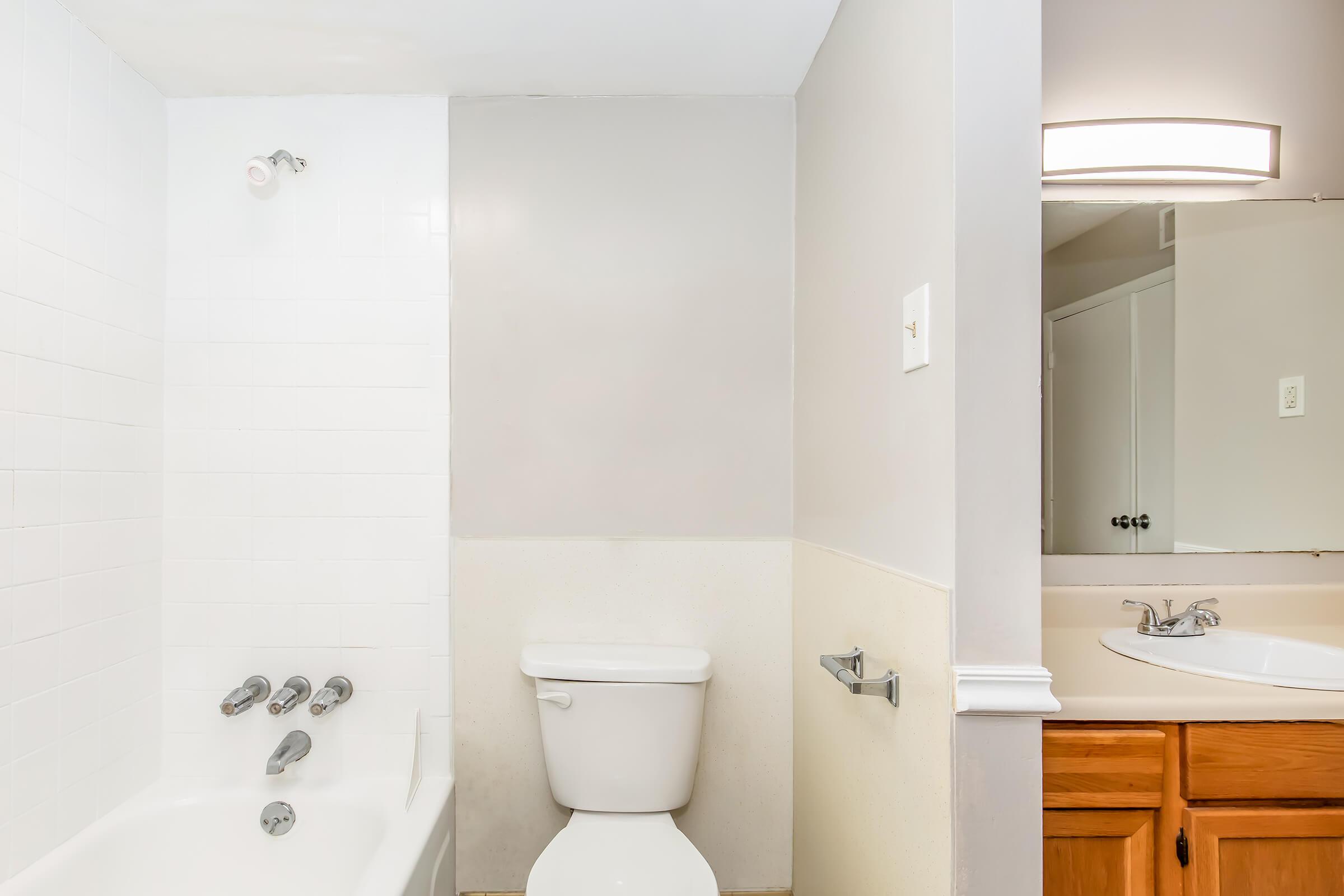A clean, well-lit bathroom featuring a white bathtub with a chrome faucet and showerhead, a white toilet, and a sink with a mirror above. The walls are a light gray color, and there is wooden cabinetry under the sink. The space is tidy and minimalistic, with bright lighting.