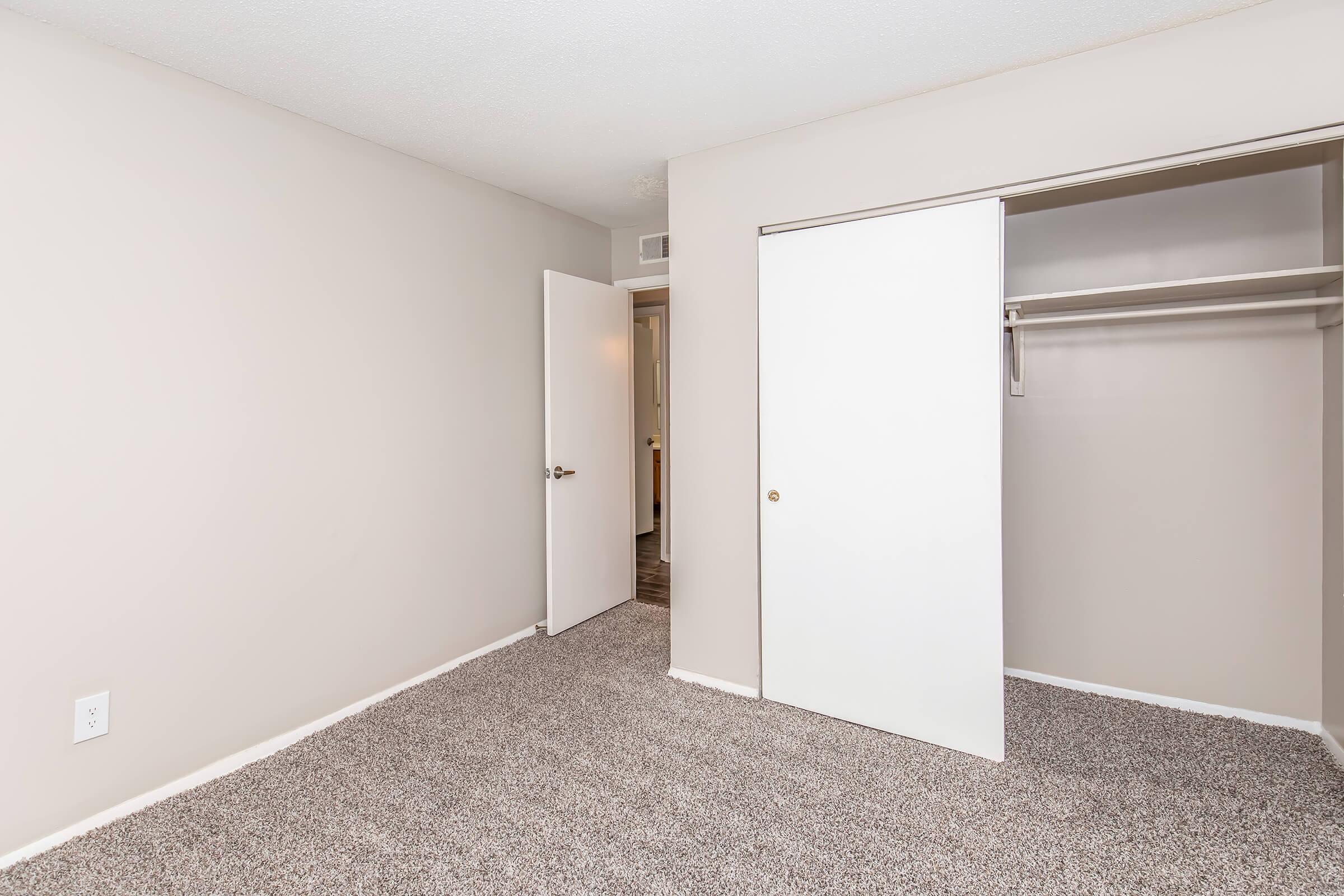 A spacious, light-colored bedroom with beige walls and gray carpet. The room features a closet with sliding white doors and an adjacent white door leading to another room. The overall atmosphere is clean and simple, ideal for personalization. Natural light is minimal, giving a cozy feel.