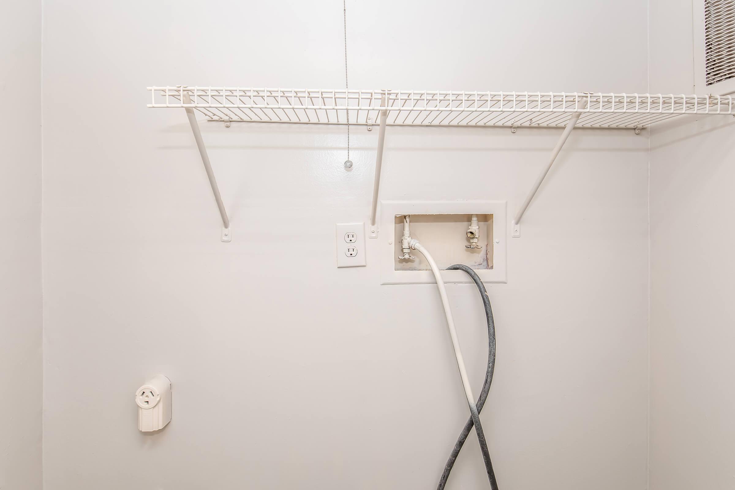 A small, empty laundry room with white walls. It features a wire shelf mounted on the wall and a wall-mounted plumbing connection for a washing machine. A power outlet is visible nearby, along with a white cord. The space appears clean and ready for installation of laundry appliances.