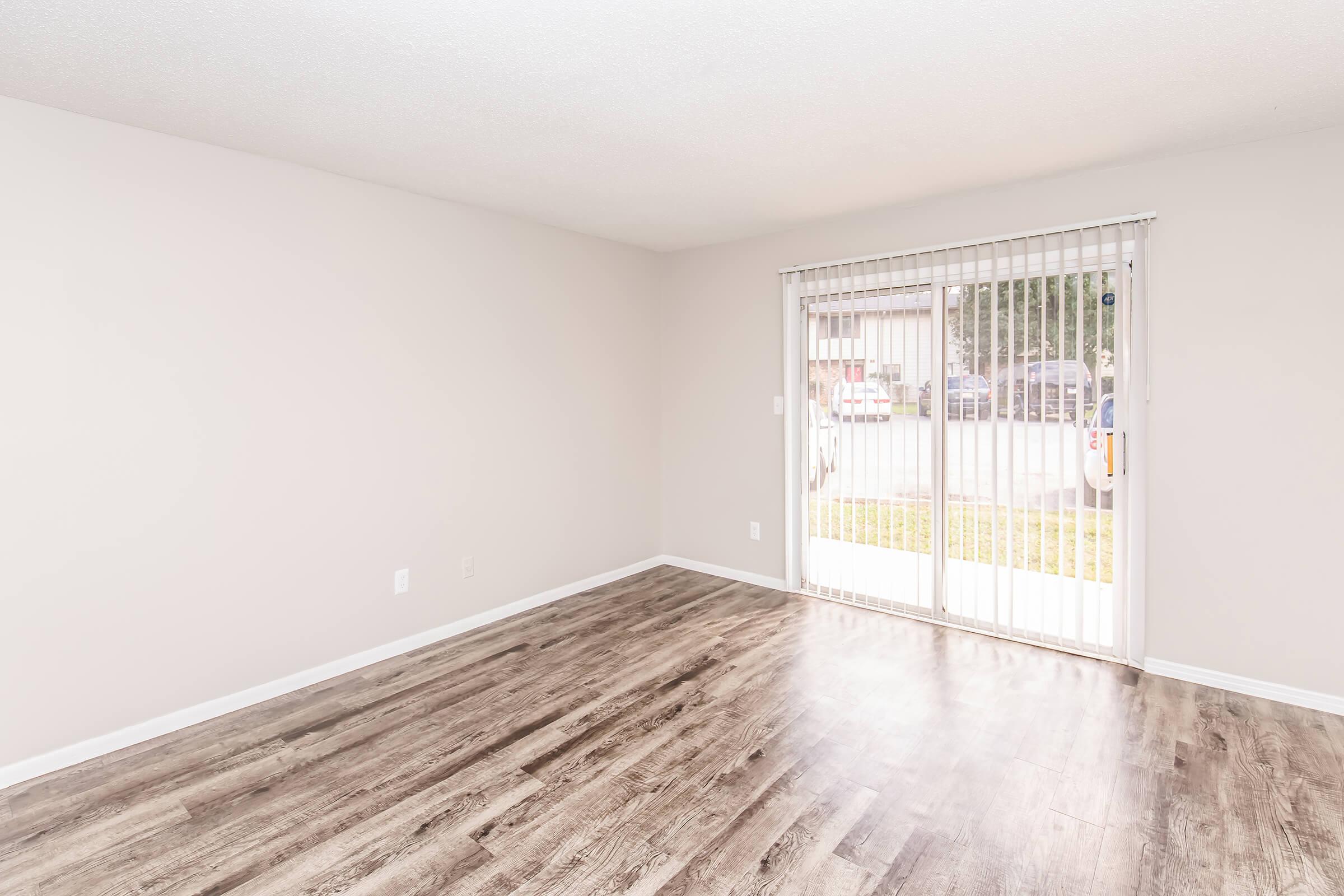 Empty interior of a room with light-colored walls and a wood-like floor. A large sliding glass door with vertical blinds allows natural light to enter, revealing a view of the outside area. The space is unfurnished and appears clean and simple, ideal for personal decoration.