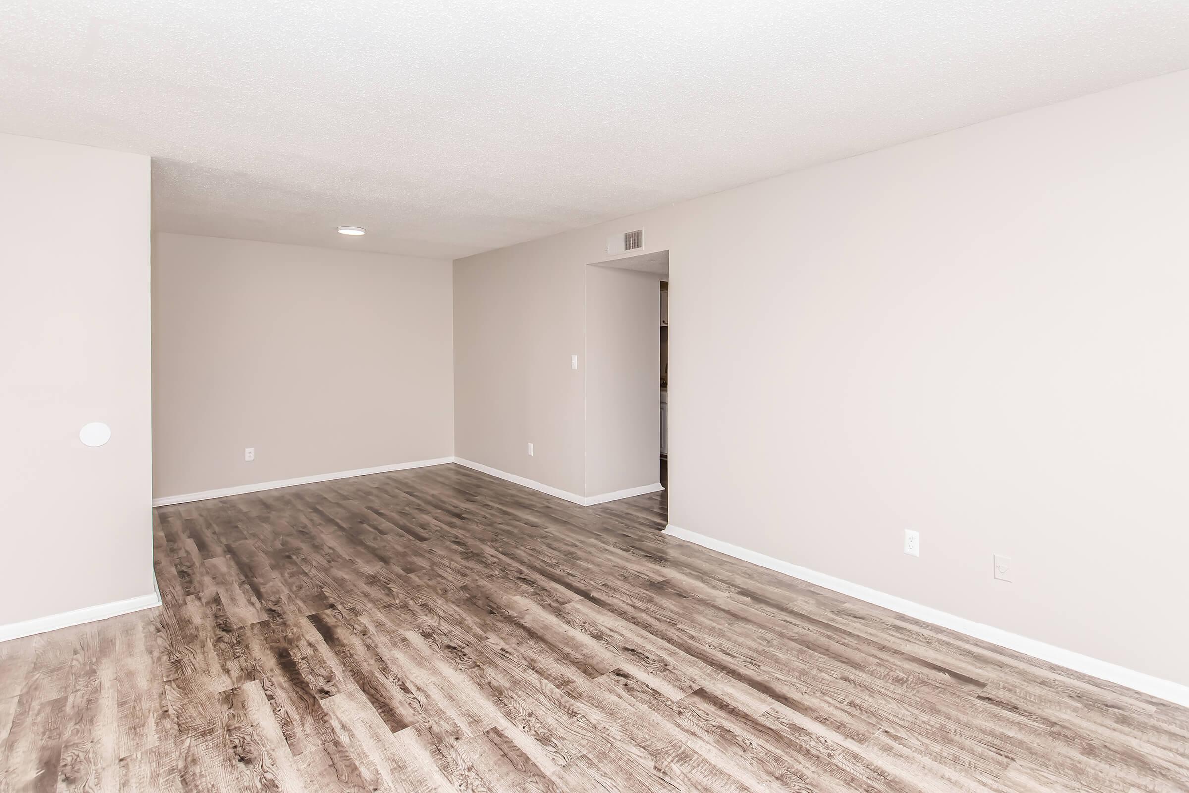 Bright, empty living space featuring light-colored walls and wooden laminate flooring. The room is well-lit with a ceiling light and has an open layout, leading to a doorway in the background. Suitable for various interior design styles.
