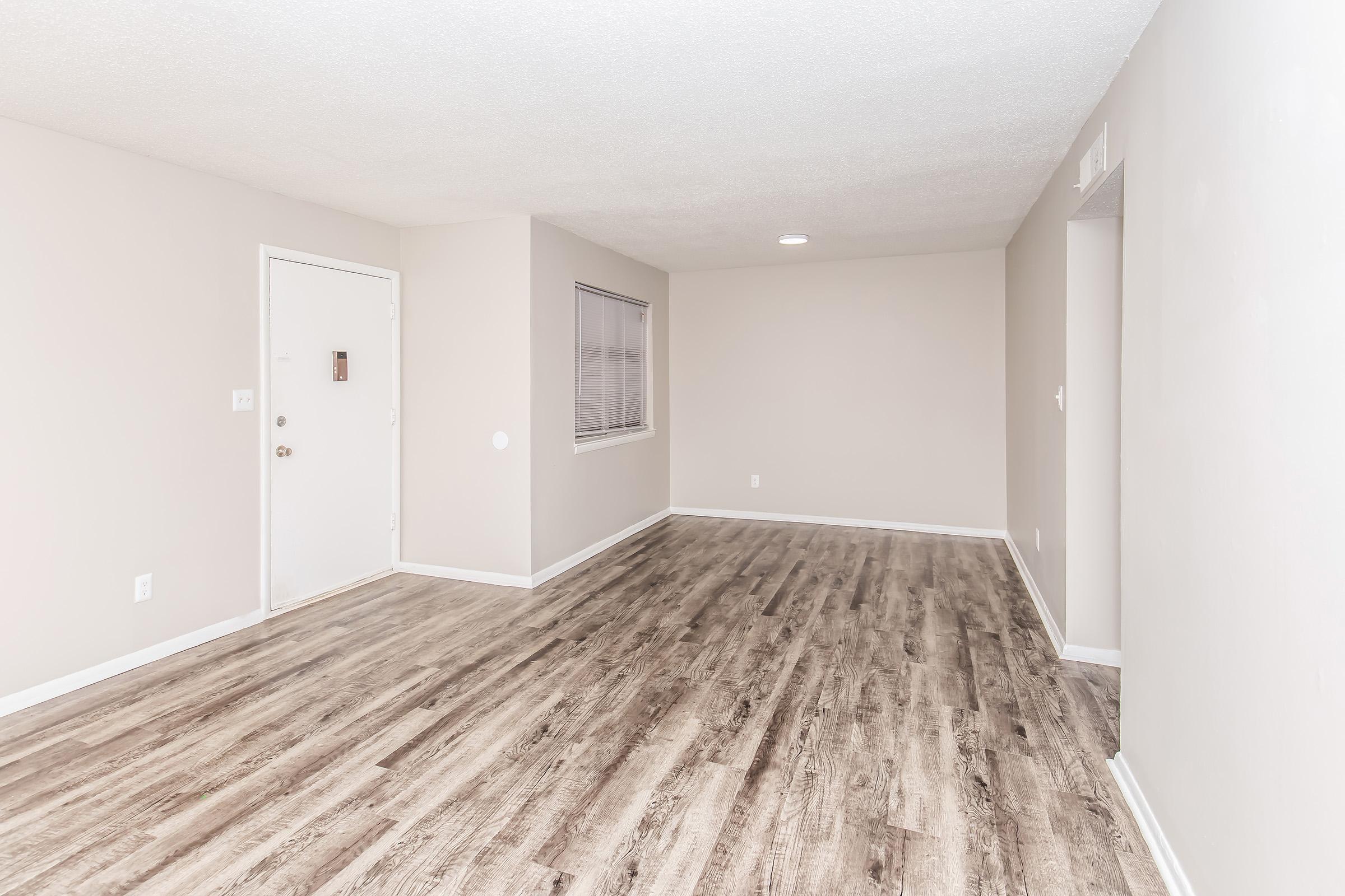A spacious, empty room with light-colored walls and a textured ceiling. The floor features wooden laminate, and there is a door on the left side leading outside. A window with blinds is visible on the right wall, and the space is well-lit with a ceiling light but lacks furniture.
