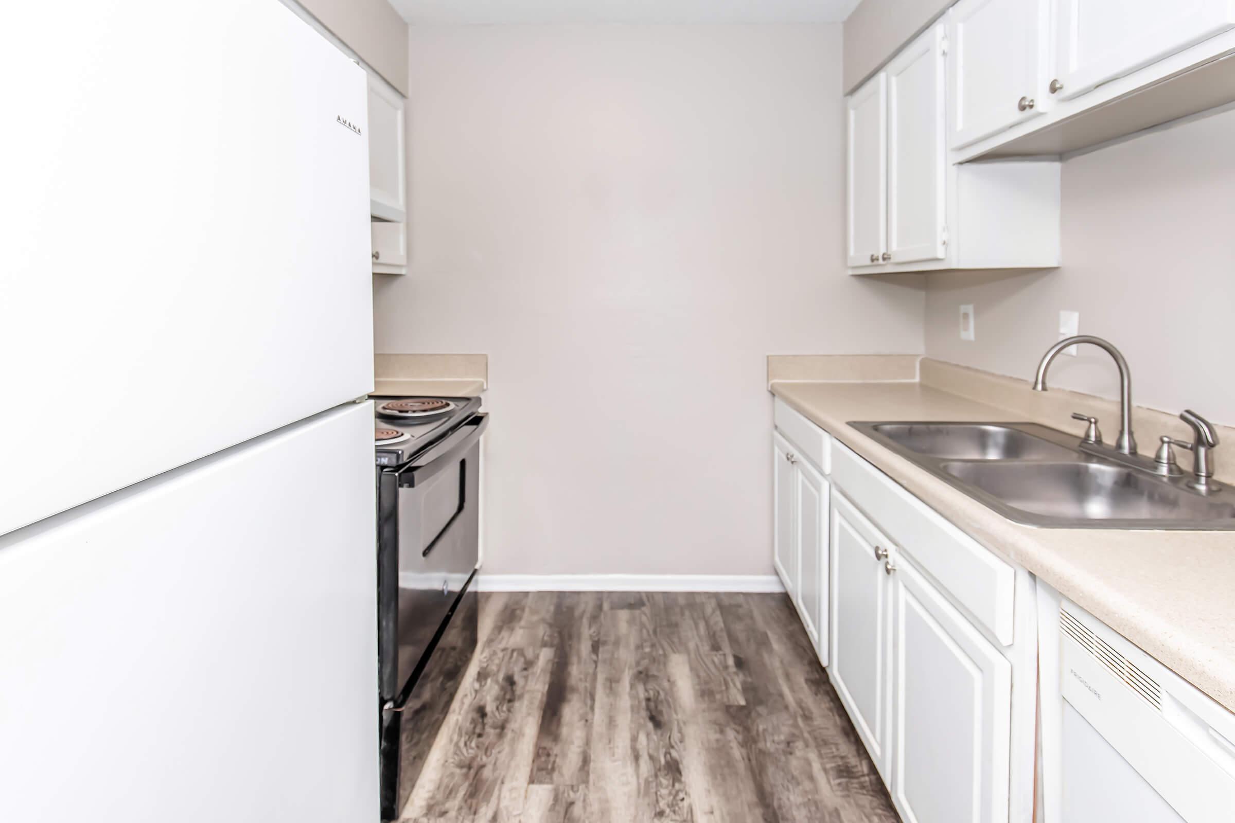 A compact kitchen featuring white cabinets, a large white refrigerator, a black stove, and dual stainless steel sinks. The countertops are light-colored, and the flooring has a wood-like appearance. The walls are painted a neutral shade, creating a bright and clean atmosphere.