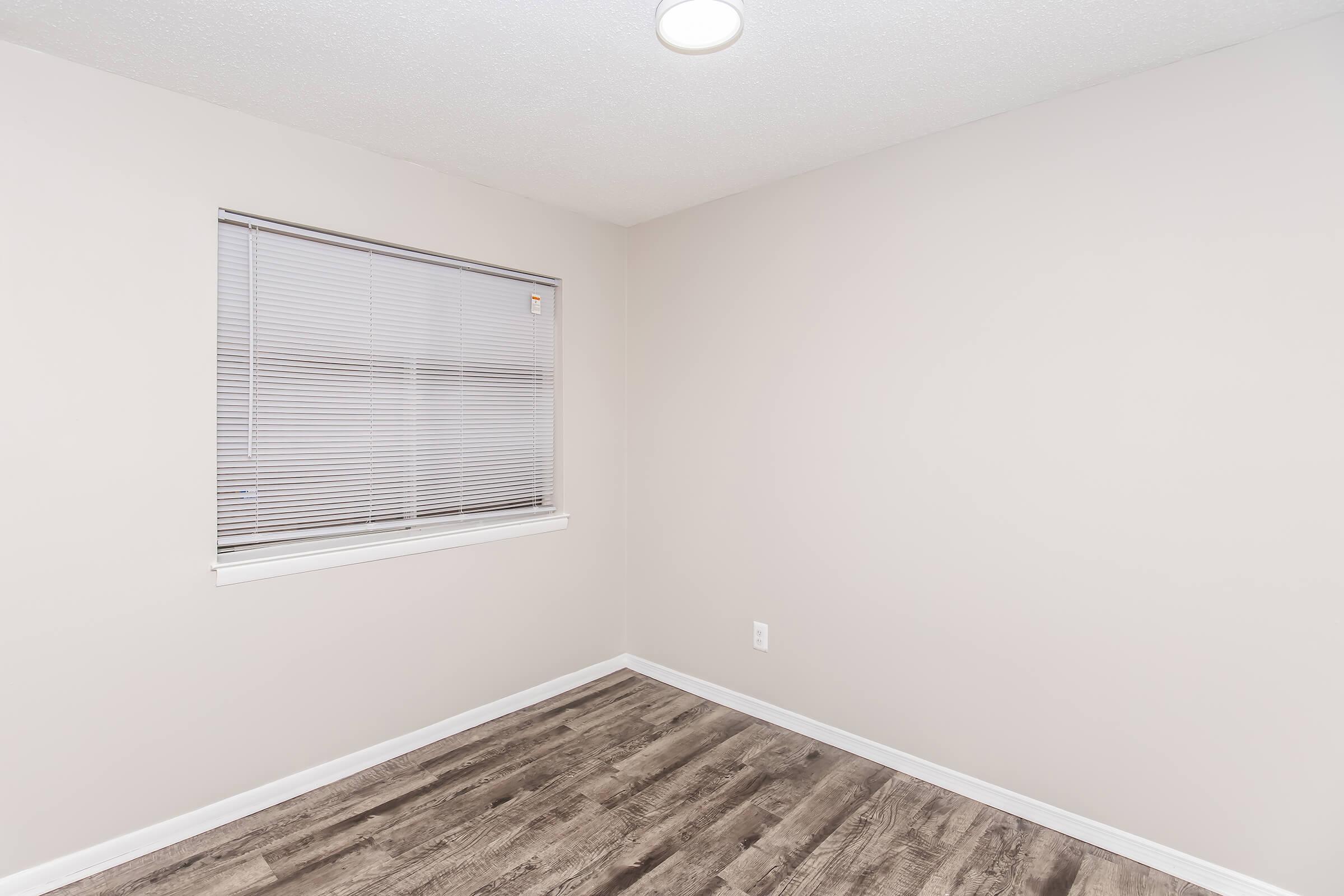 Empty room with light-colored walls and a window featuring closed blinds. The floor is covered with wood-like laminate flooring, and there is a ceiling light fixture visible. The overall ambiance is bright and minimalistic.