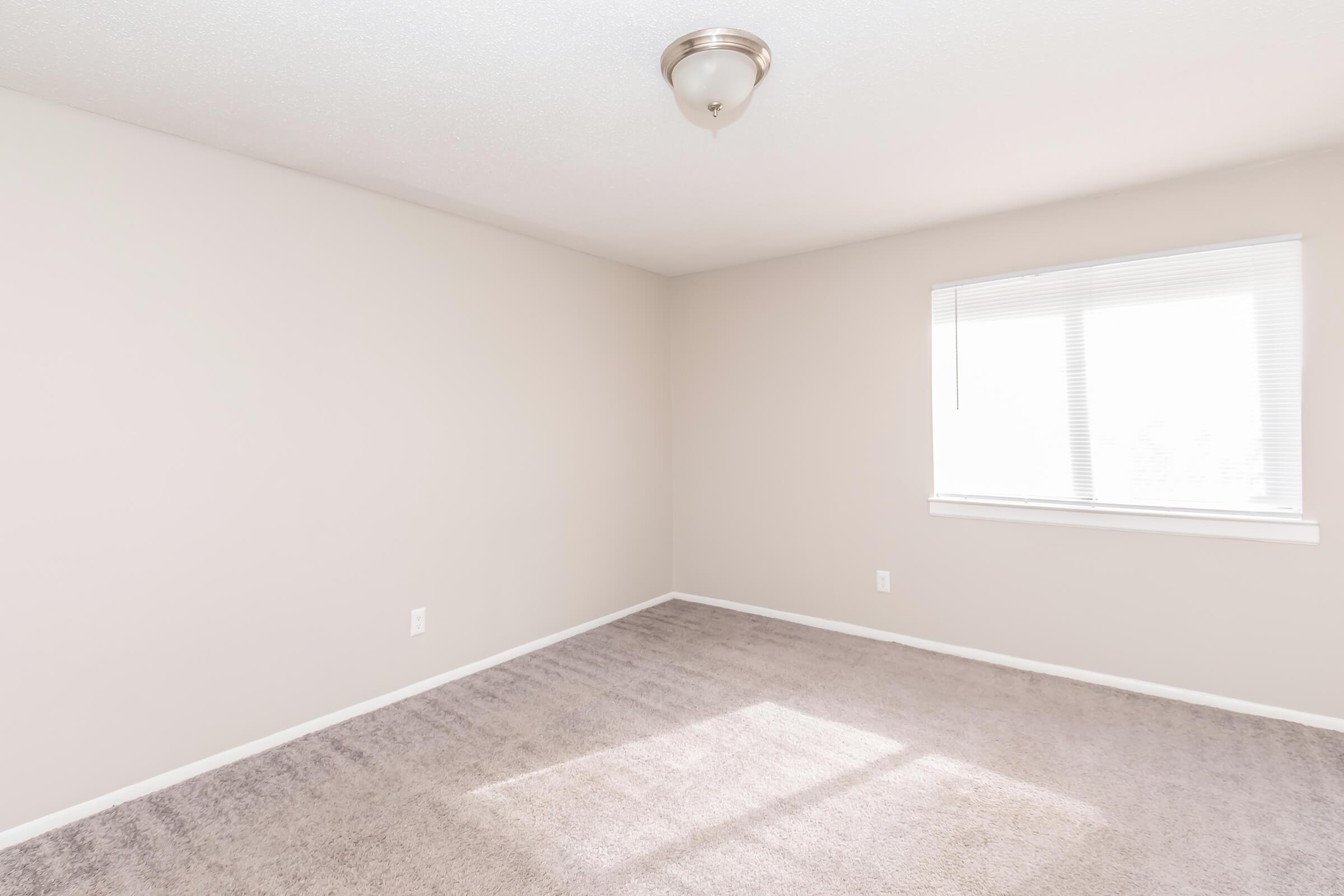 A vacant room with light beige walls, a light brown carpet, and a window with blinds allowing natural light. The ceiling features a simple light fixture, and the space is minimally decorated, giving it a spacious, clean appearance.