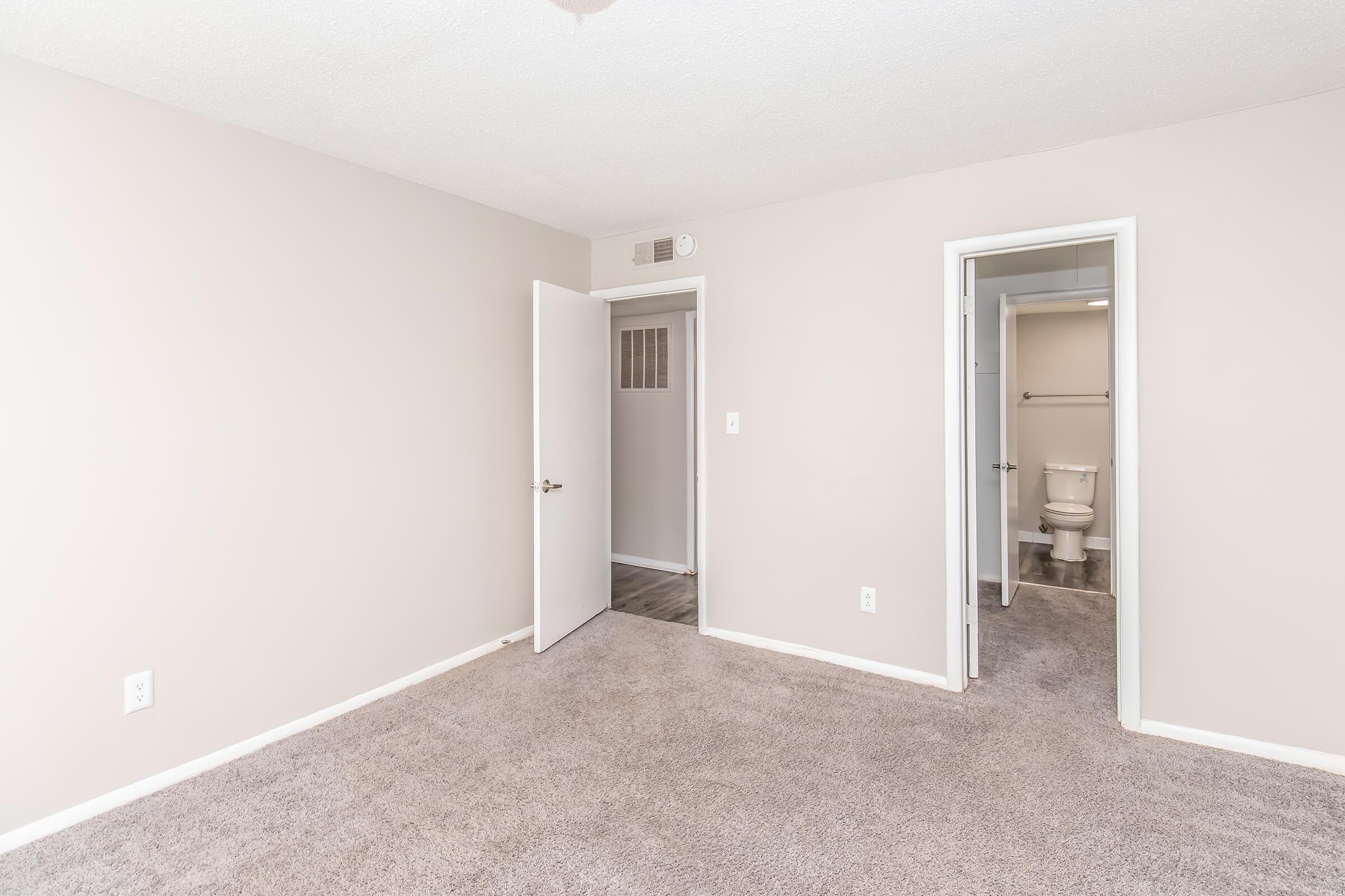 A spacious, unfurnished room with light beige walls and carpeted flooring. One door leads to a small bathroom, visible in the background, while another door is partially open. The overall atmosphere is bright and simple, suggesting a clean and modern living space.