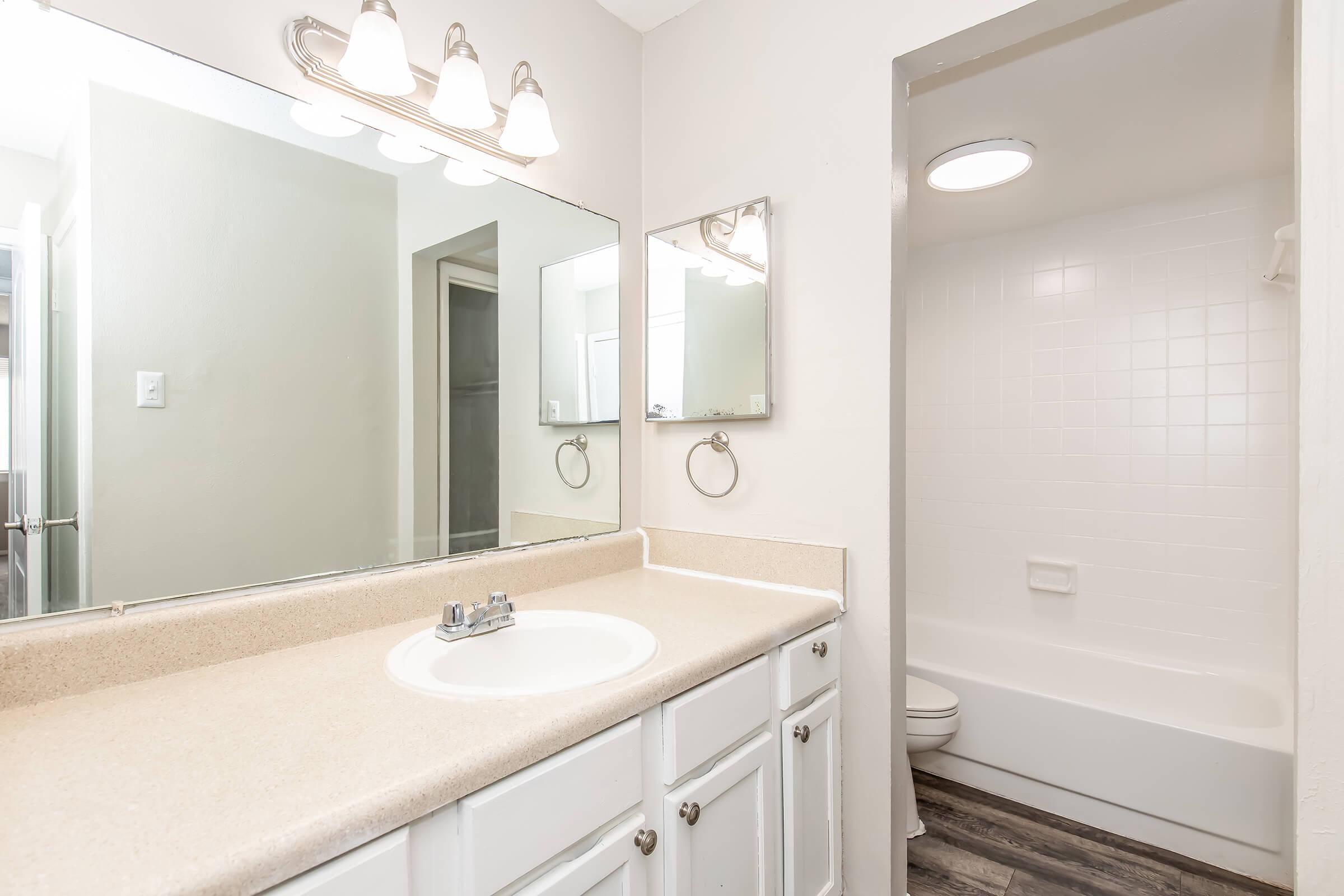 A modern bathroom featuring a double-sink vanity with a light-colored countertop, three wall-mounted light fixtures above a large mirror. To the right, a separate area with a bathtub and shower, white tiled walls, and a circular ceiling light. Neutral-colored walls and flooring complete the clean, contemporary look.