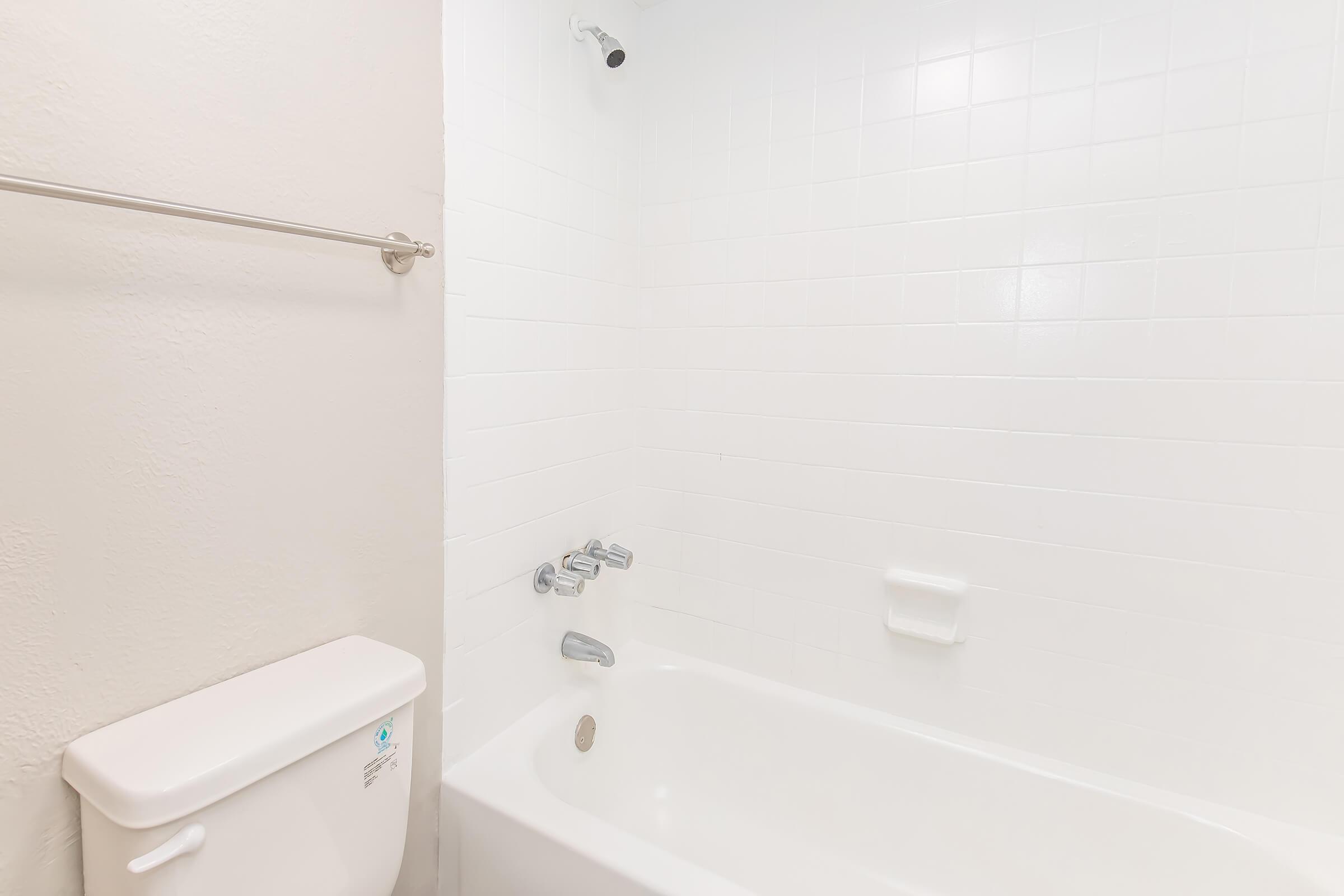 A clean, minimalistic bathroom featuring a white tiled wall, a bathtub, a wall-mounted faucet, and a toilet. The space is well-lit with a bright, white appearance, creating a fresh and open atmosphere. A towel rack is mounted on the wall, completing the simple design.