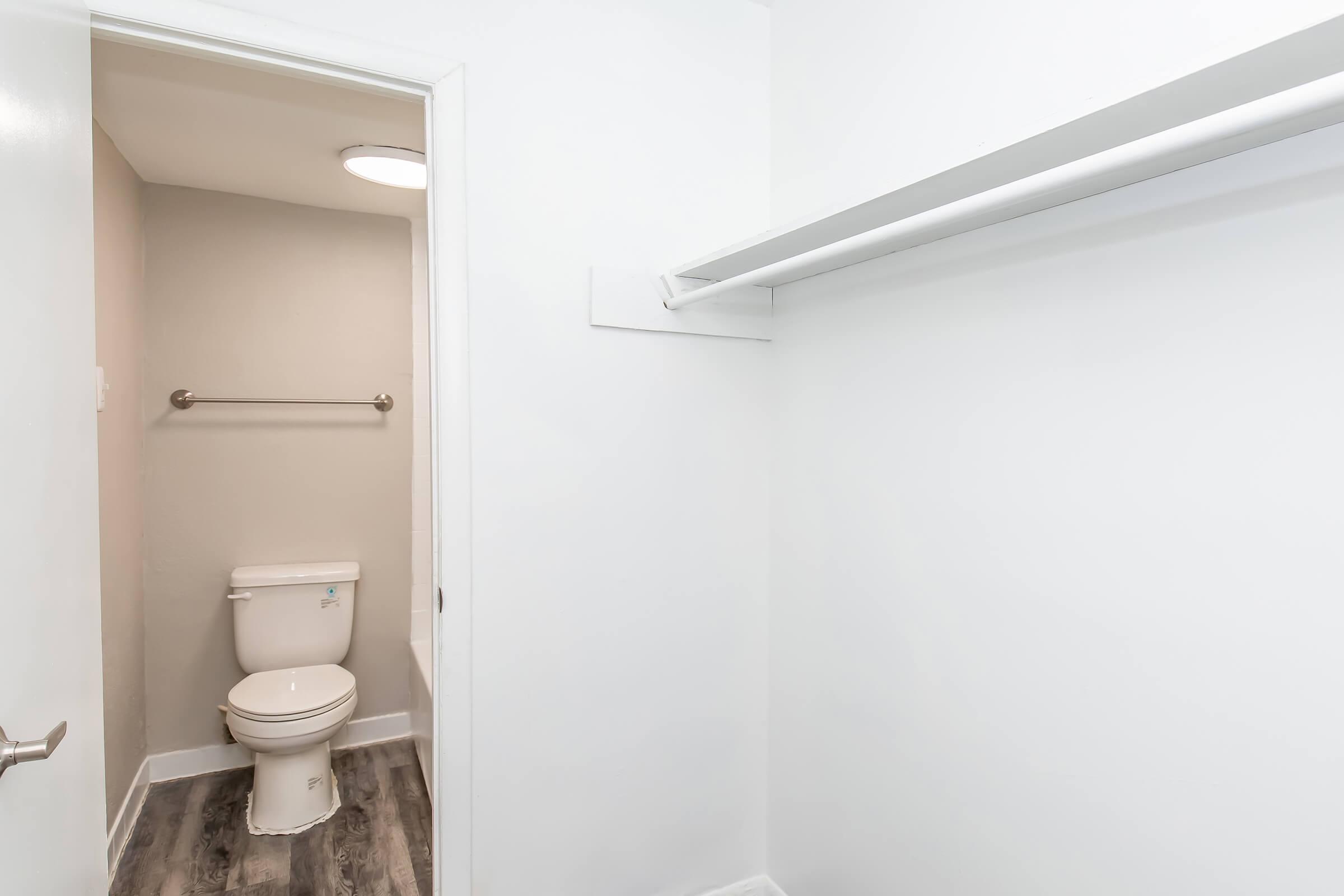 Interior view of a small bathroom featuring a white toilet, a light gray wall, and a wall-mounted towel bar. The space includes a door leading to another room and has a simple, clean design with light-colored flooring. A single overhead light fixture illuminates the area.