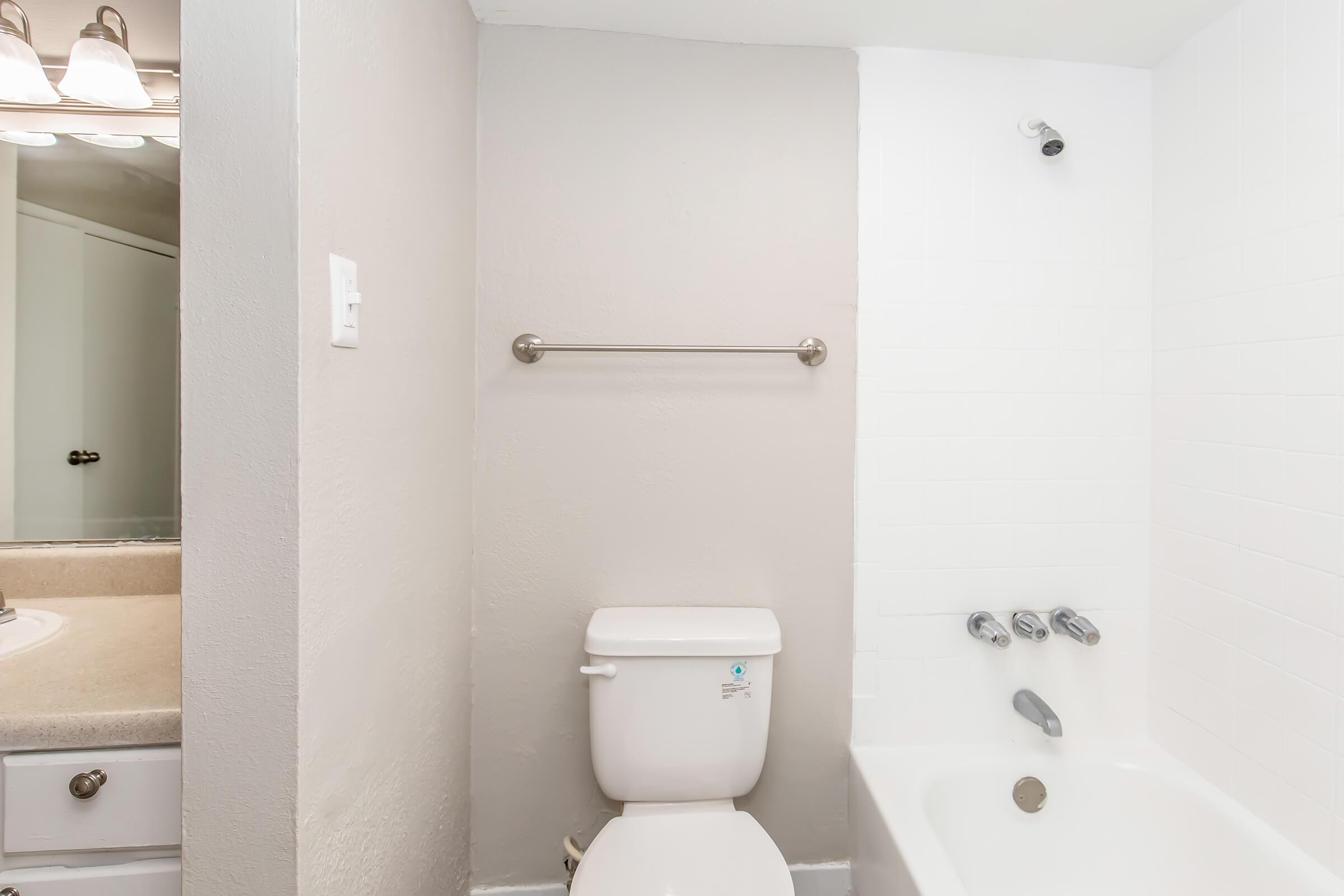 A clean, modern bathroom featuring a white bathtub, a wall-mounted towel bar, and a toilet. The wall is painted in soft gray, and there's a light fixture visible in the background. The space has a minimalistic design with a neutral color palette.