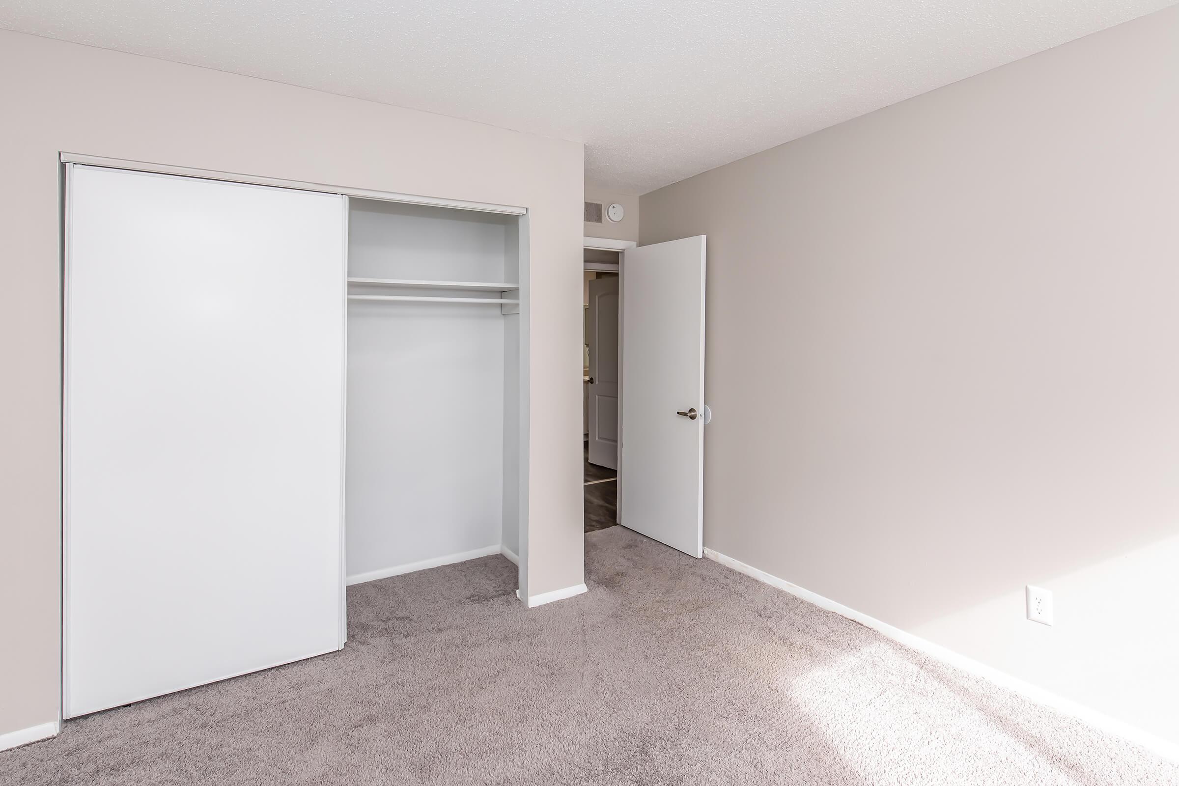 Empty interior of a room featuring light-colored walls and carpet. There is a closed white sliding door to a closet on the left and a white door leading to another area on the right. Natural light illuminates the space, creating a bright and airy atmosphere.