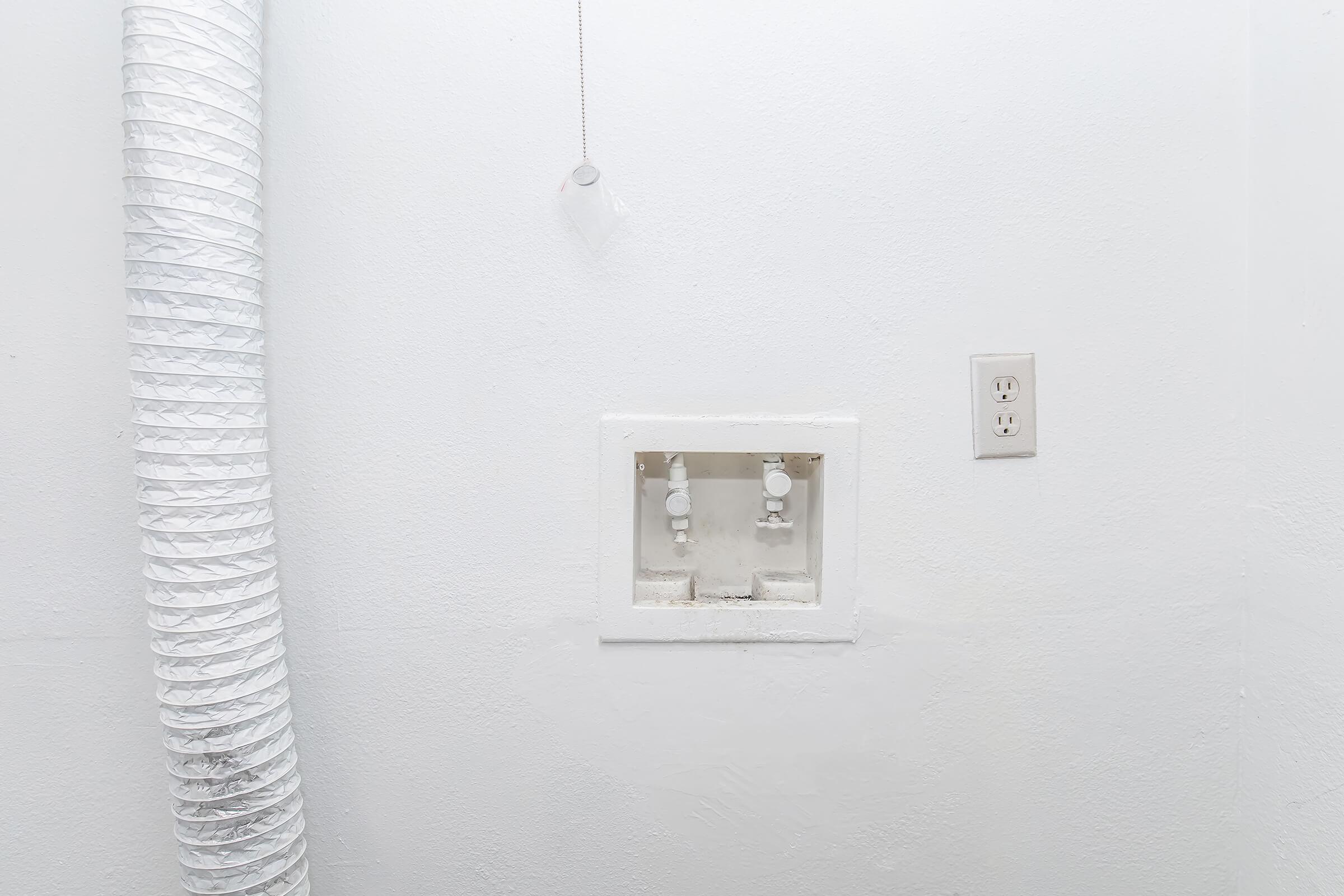 A close-up of a white wall with two water valves mounted in a rectangular box, adjacent to a standard electrical outlet. A flexible, white duct is visible, extending from the left side of the image, contributing to a minimalistic and functional aesthetic. The overall color scheme is predominantly white.
