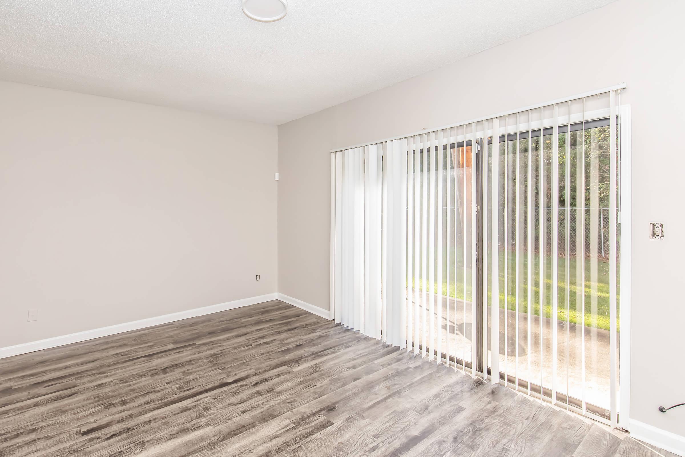 A bright and empty room with light-colored walls and wood-like flooring. There is a sliding glass door with vertical blinds leading to an outdoor area. Natural light filters in, creating an open and airy atmosphere. The space is well-lit and inviting, perfect for customization.