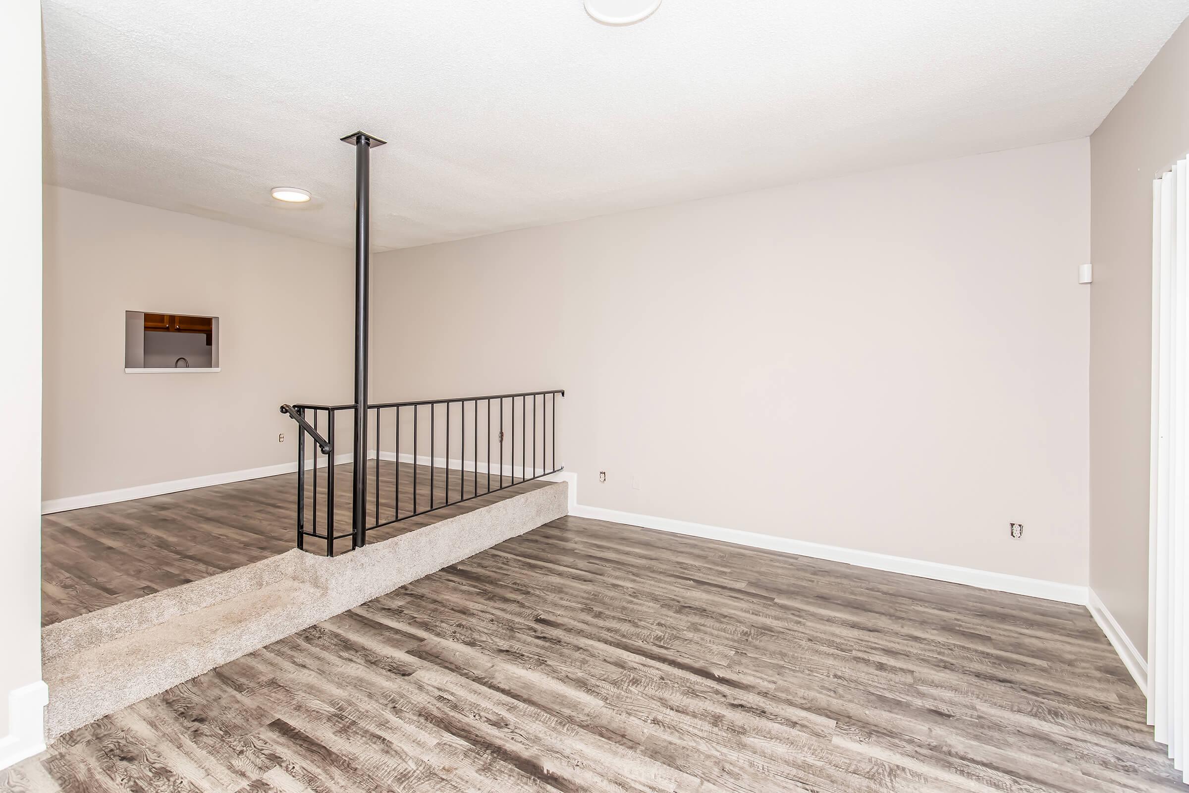 Spacious interior of a room with light-colored walls and new flooring. Features a staircase with a black railing leading to a raised area. Natural light enters through a small window, creating a bright and inviting atmosphere. The room is empty, providing a blank canvas for personal decoration.