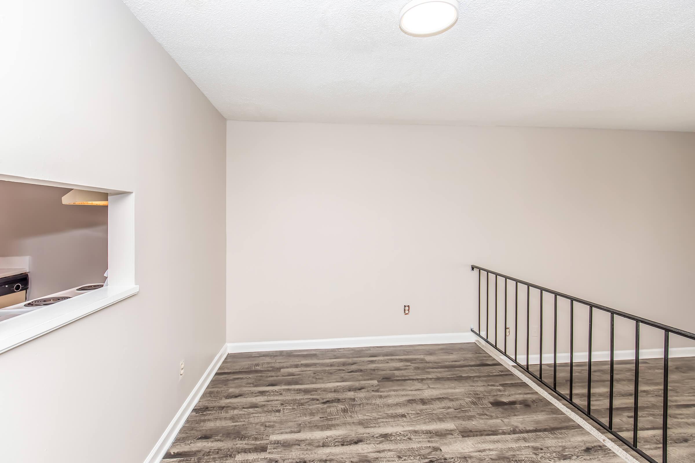 An empty room with light-colored walls and a wooden floor. There is a window opening to a kitchen area visible in the background. A black railing runs along one side, and a ceiling light illuminates the space. The atmosphere is bright and minimalist, creating a spacious feel.