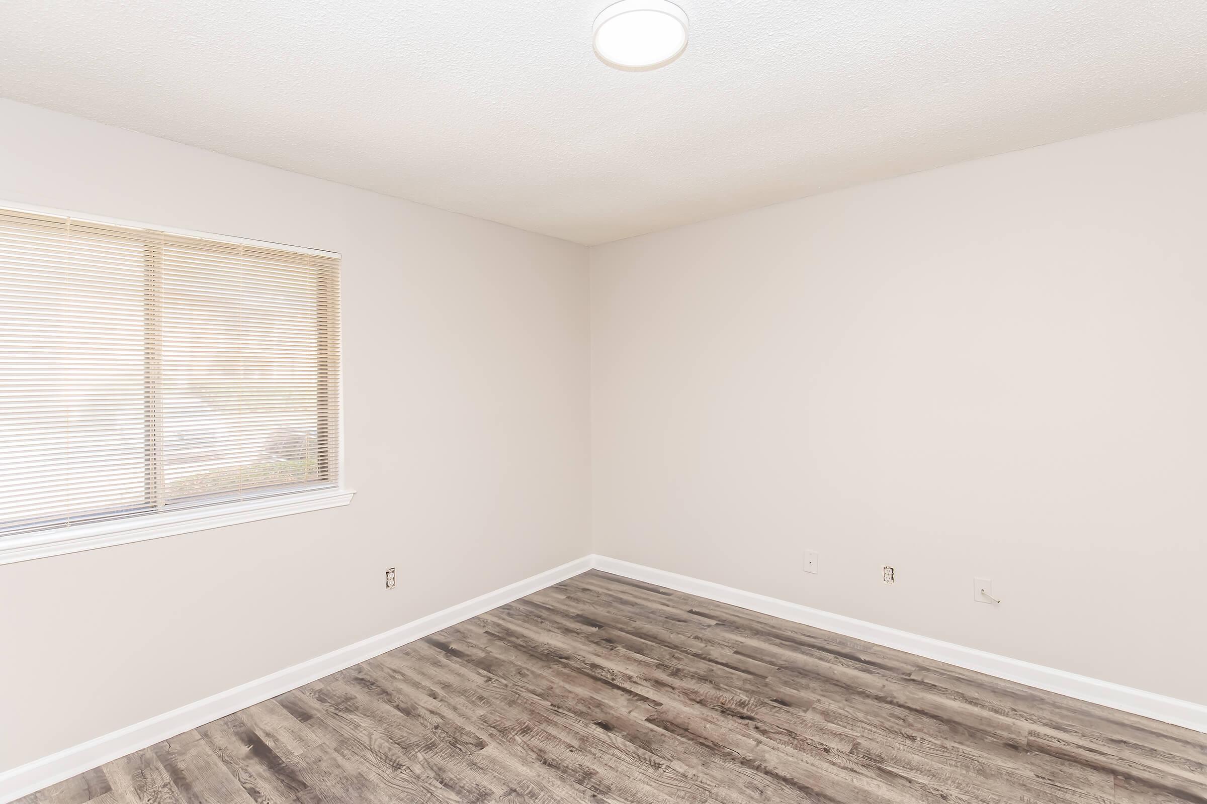 A spacious, empty room with light-colored walls and a large window covered by blinds. The floor features light brown, wood-like laminate. A ceiling light fixture provides illumination, and there are no furnishings present, creating a clean and minimalistic aesthetic.