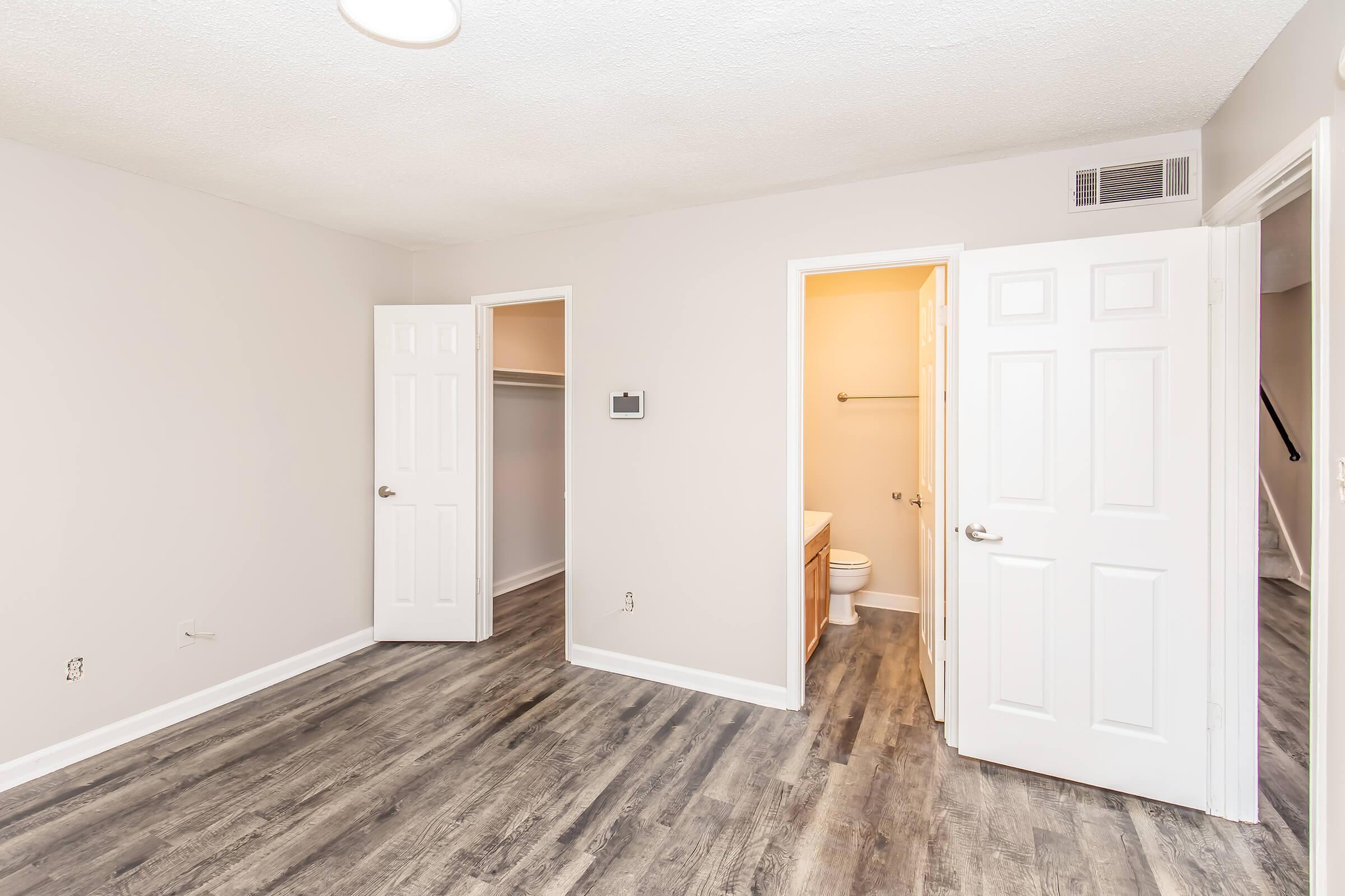 A spacious, empty room featuring light-colored walls and laminate flooring. There are two open doors leading to closets, a bathroom visible through one door, and a hallway beyond. The ceiling has a circular light fixture, providing ample lighting.