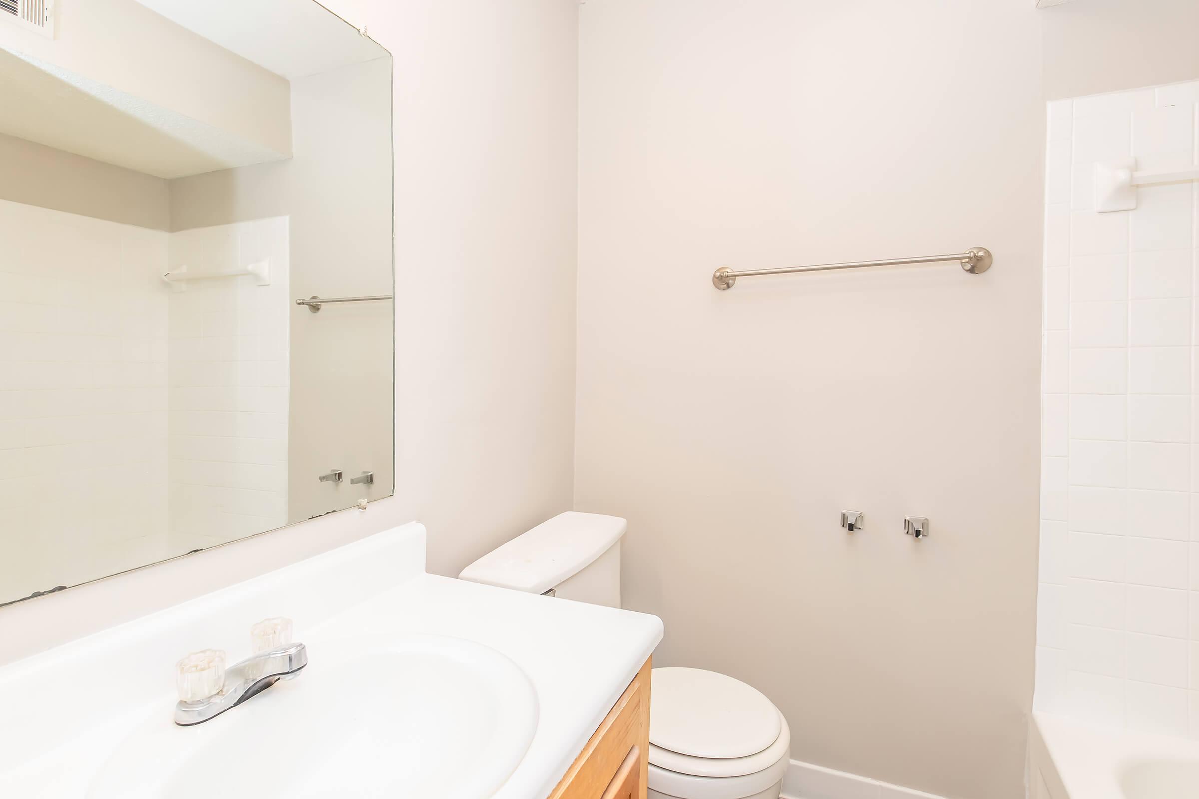 A clean, modern bathroom featuring a white toilet, a sink with a single faucet, and a large mirror above the sink. The walls are painted light beige, and there is a towel bar mounted on the wall. A bathtub is visible on the right side, and the overall ambiance is bright and uncluttered.
