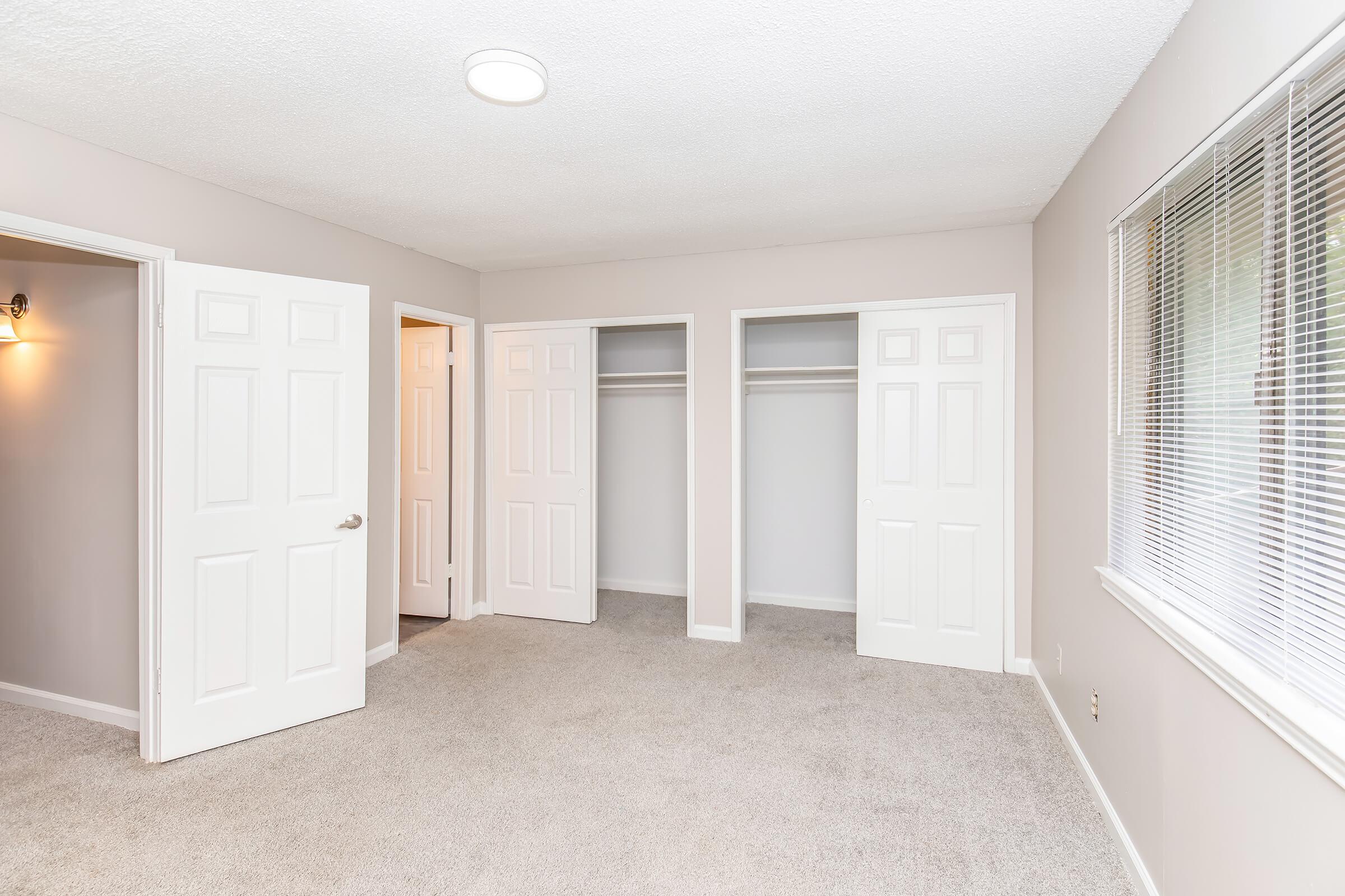 Bright and spacious bedroom featuring light-colored walls, plush carpet flooring, and two large closets with double doors. Natural light floods in through a window with blinds, and there is a ceiling light fixture. Adjacent doorways lead to additional rooms or spaces.