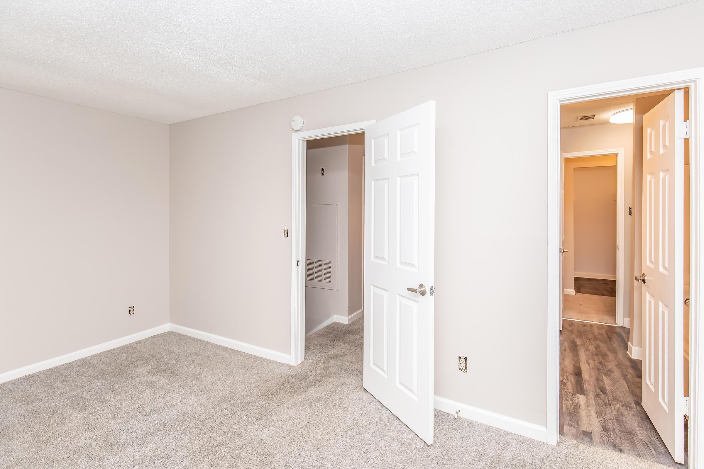 A spacious, empty room with light gray carpeting and neutral-colored walls. One door is open, leading to another room, while another door connects to a closet or utility area. Soft natural light fills the space, creating a welcoming atmosphere.