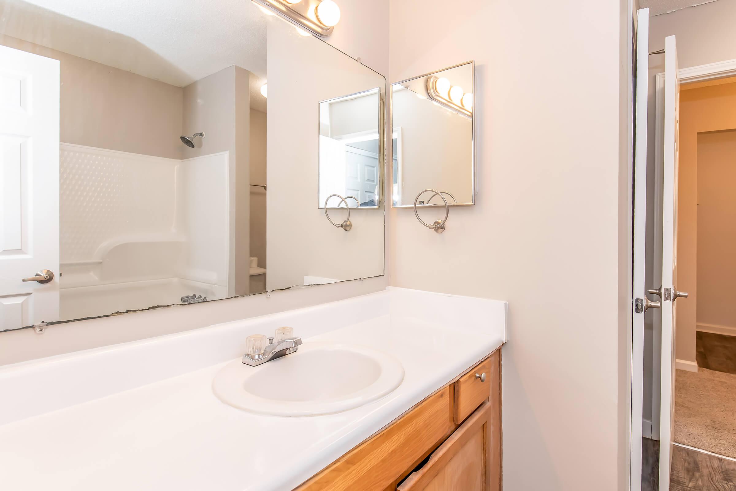 A clean and modern bathroom featuring a white countertop with a sink, two mirrors above, and wooden cabinetry. The bathroom has a tub with a showerhead, light gray walls, and a door leading to another room, creating a bright and spacious atmosphere.