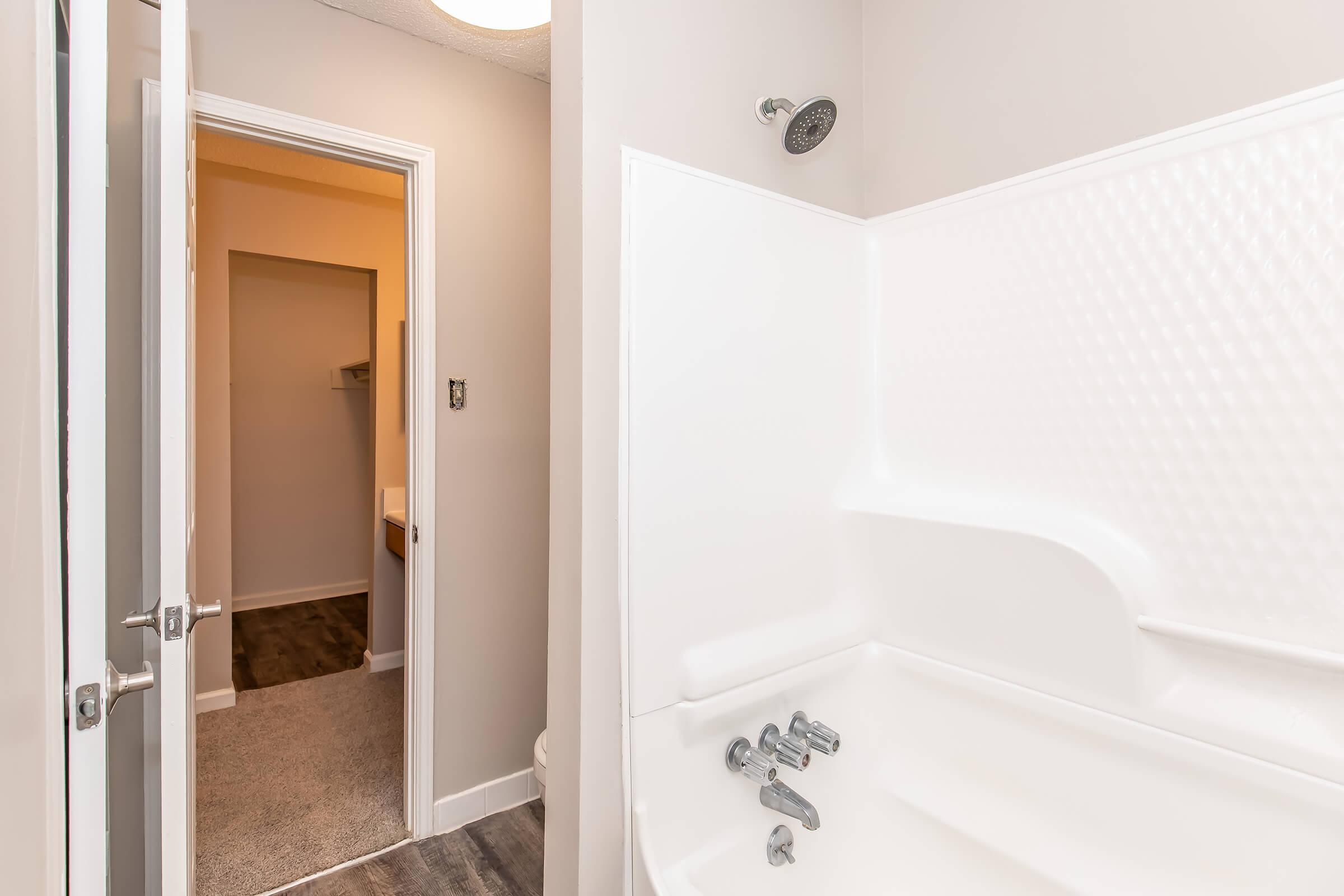 A bathroom with a bathtub and showerhead, featuring a light-colored wall and a white tub. The doorway leads to another room beyond, with warm lighting visible. The floor has a dark finish, contrasting with the light walls and tub.