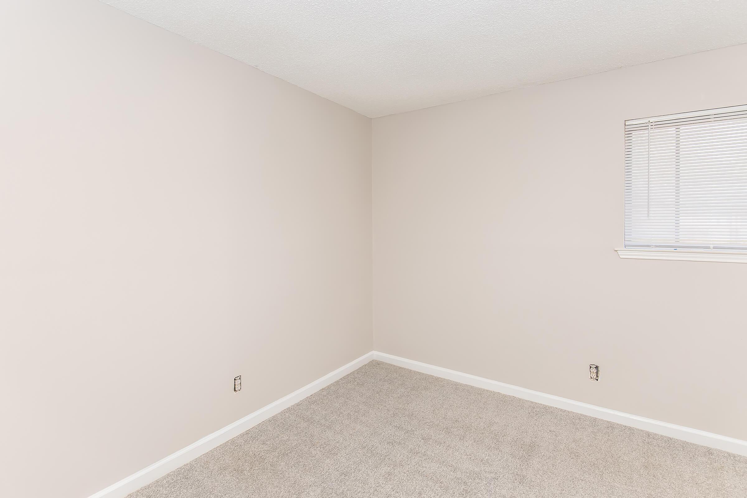 Empty room with light beige walls and a small window with vertical blinds. The floor is covered in gray carpet. The space appears clean and unoccupied, featuring two electrical outlets on the walls. Natural light enters through the window, enhancing the neutral color scheme.
