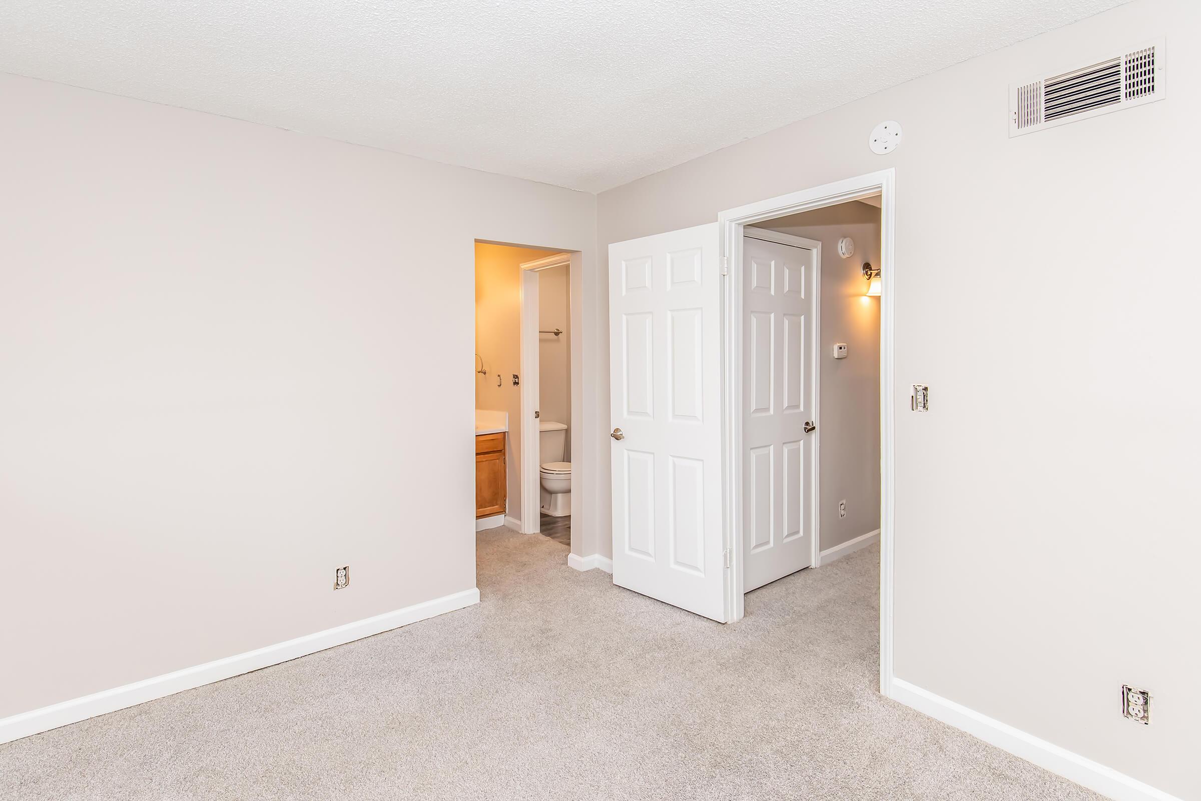 Empty room featuring light beige walls and carpet. A doorway leads to a bathroom with light fixtures visible. Two closed white panel doors are present, one leading to the bathroom and the other to an unknown area. The space is bright and uncluttered, typical of a rental or newly remodeled apartment.