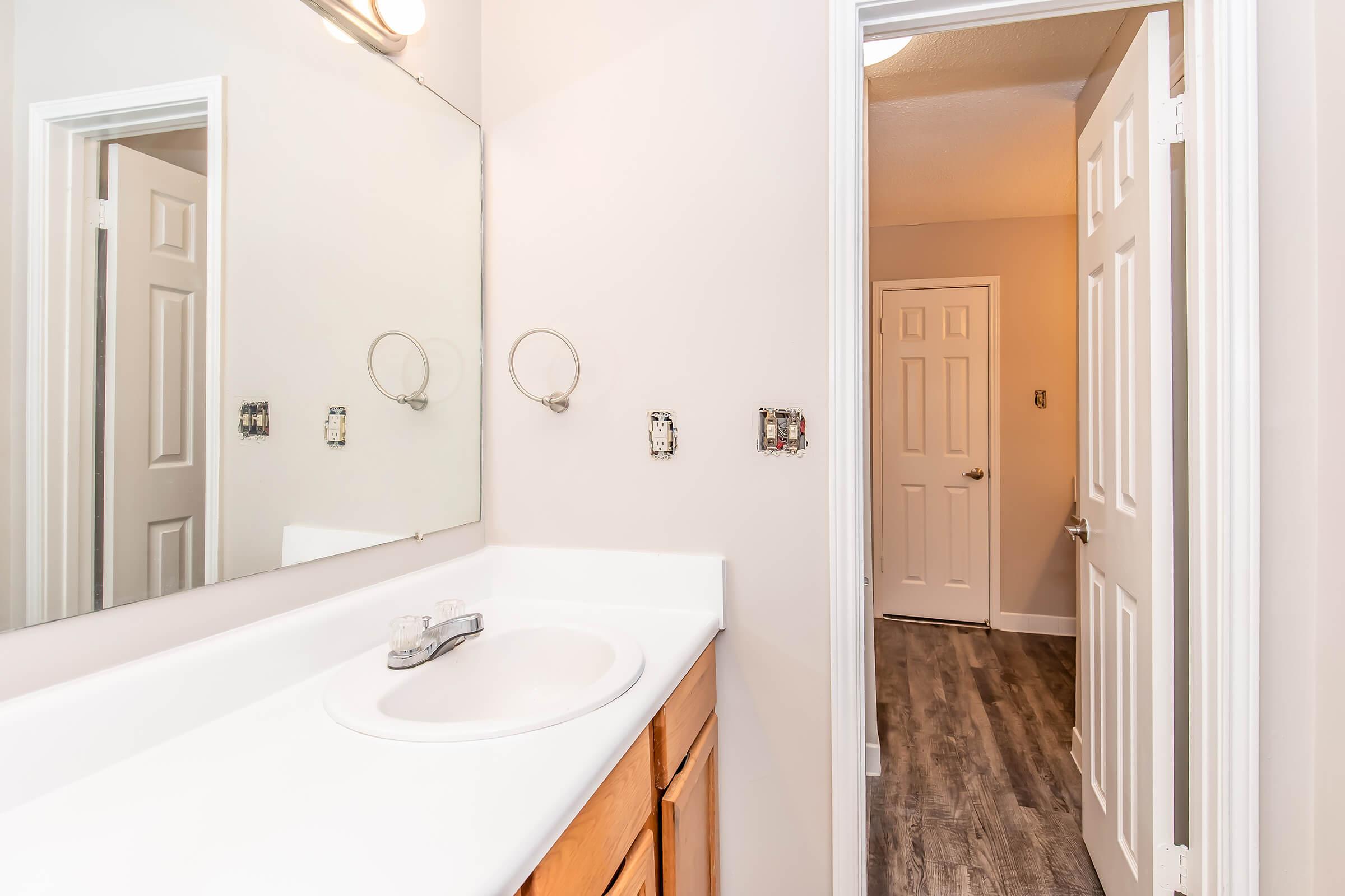 A bathroom scene featuring a white countertop with a sink, a large mirror above, and beige walls. There's wooden cabinetry below the sink, and a doorway in the background leading to another room. The flooring is a light wood style, and there are unconnected fixtures on the wall near the mirror.