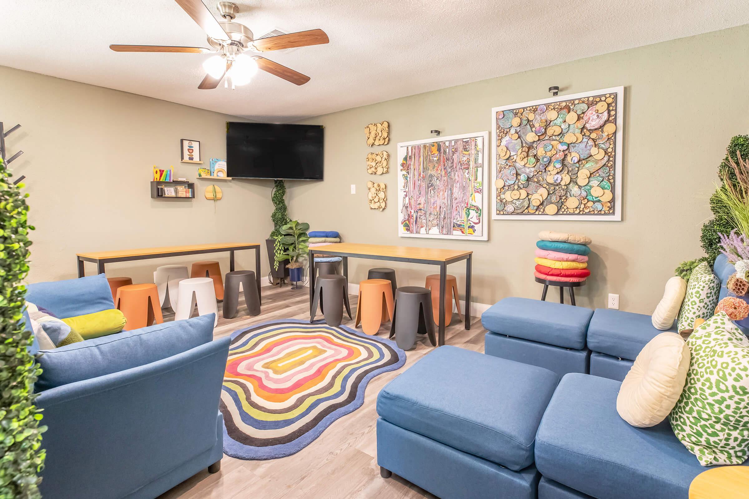 A vibrant room featuring blue sectional sofas with colorful pillows, a round patterned rug, and a mix of tables and stools. Wall art displays abstract designs, and a TV is mounted on one wall. Green plants add a fresh touch to the inviting and playful atmosphere.