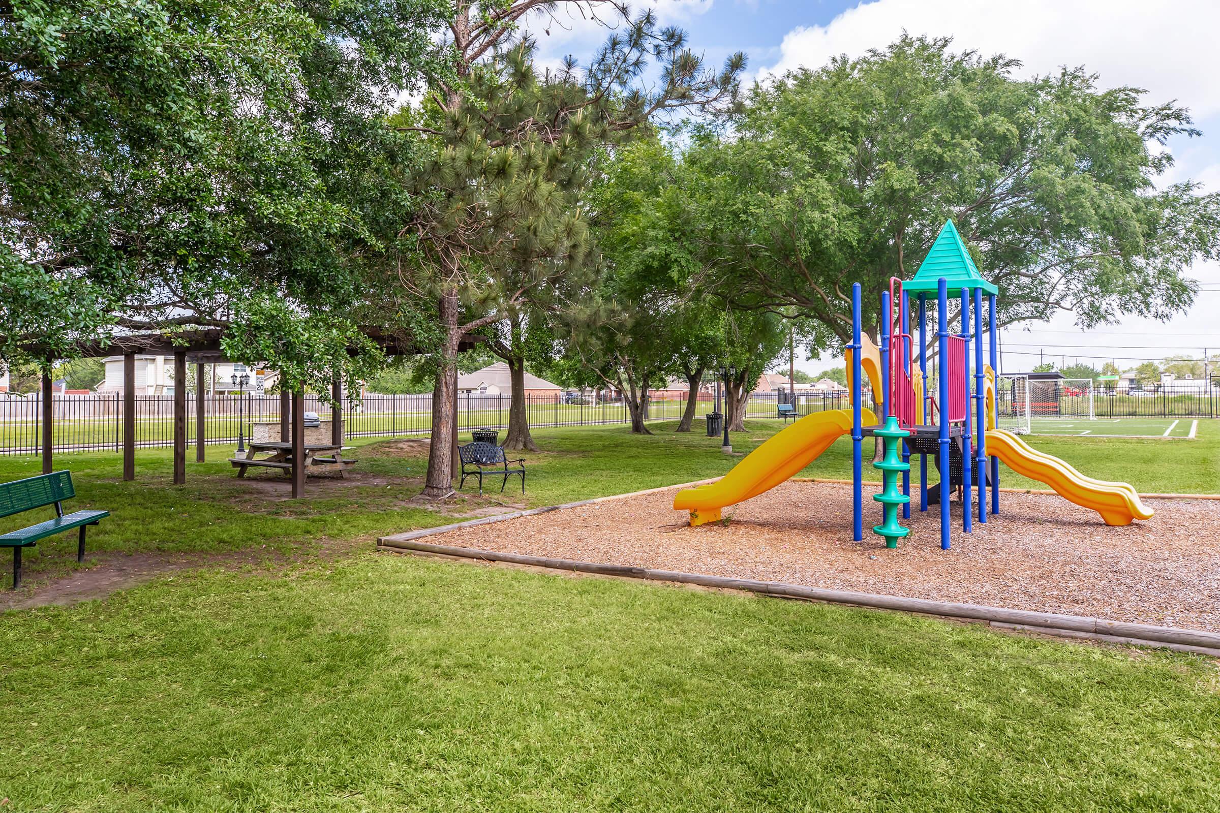 A colorful playground with yellow and orange slides sits on a bed of gravel, surrounded by grassy areas. Nearby, there are benches for seating and trees providing shade. In the background, a fenced area and faint outlines of sports fields can be seen under a partly cloudy sky.