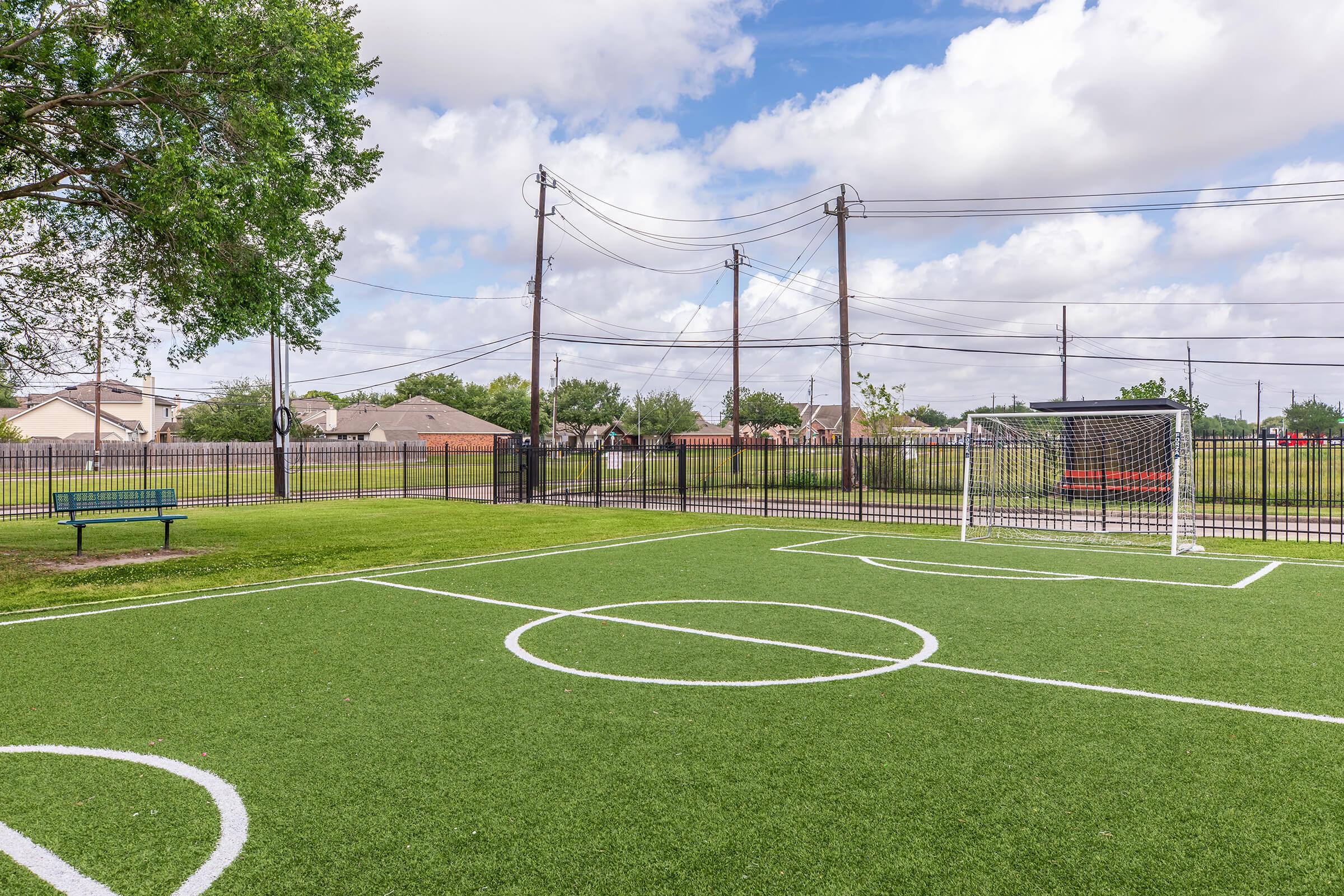 A small soccer field with artificial turf, featuring a goal at one end. There are white boundary lines marking the center circle and penalty area. In the background, a park bench and a fenced area with power lines and distant residential buildings. The sky is partly cloudy.