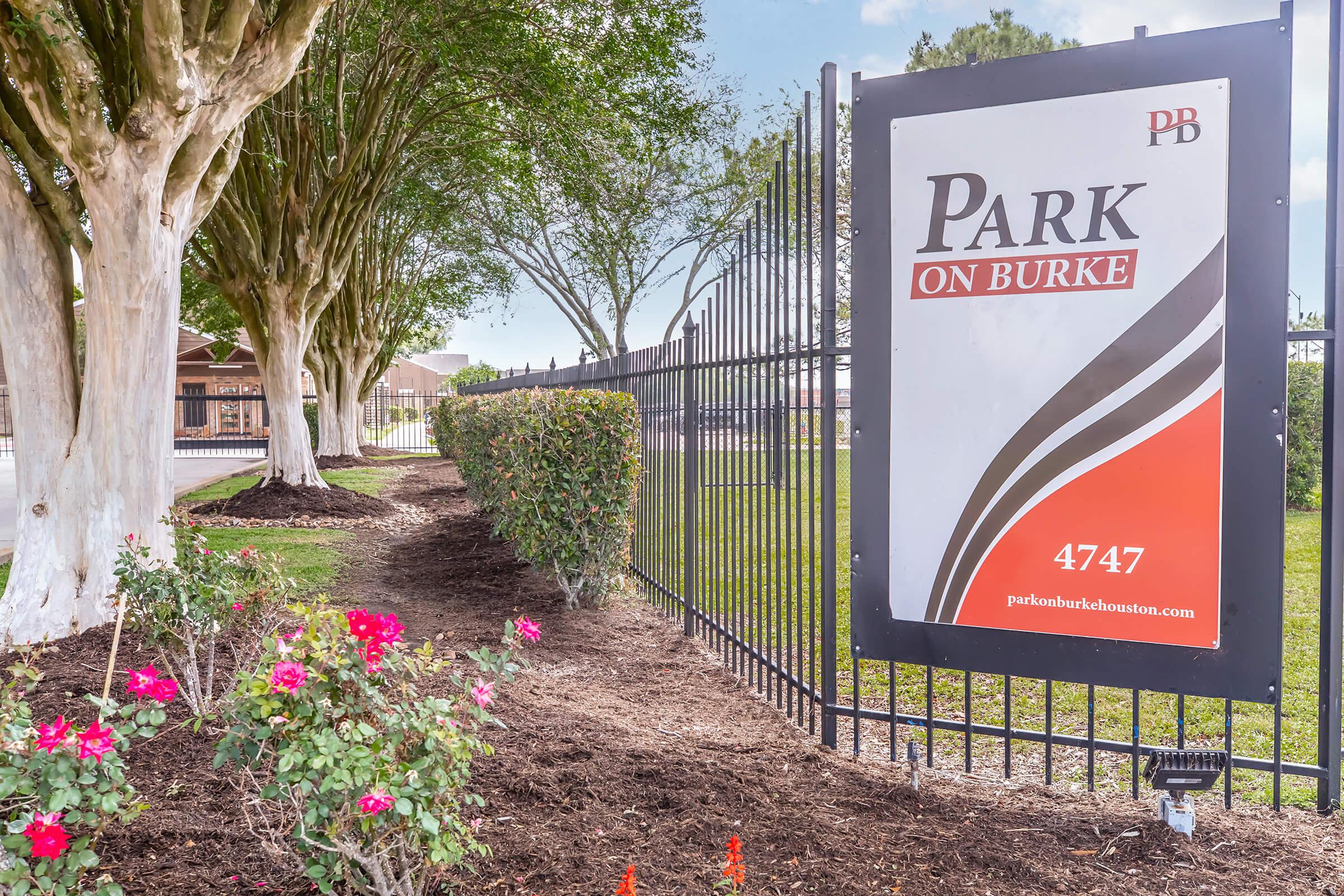 Sign for "Park on Burke" located at 4747, with a colorful design featuring red, black, and white stripes. The sign is surrounded by neatly trimmed bushes and flowers, set against a backdrop of trees and a blue sky. A fence encloses the area, indicating it is part of a residential or community space.