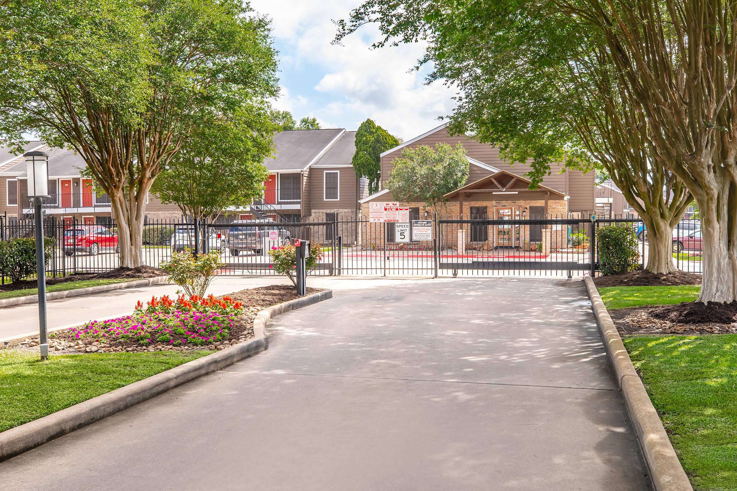 A landscaped entrance leading to an apartment complex, featuring a paved pathway, flower beds with colorful plants, and a gated area in the background. Surrounding trees provide shade, and there are buildings visible behind the gate. The scene is bright and inviting, set in a suburban environment.